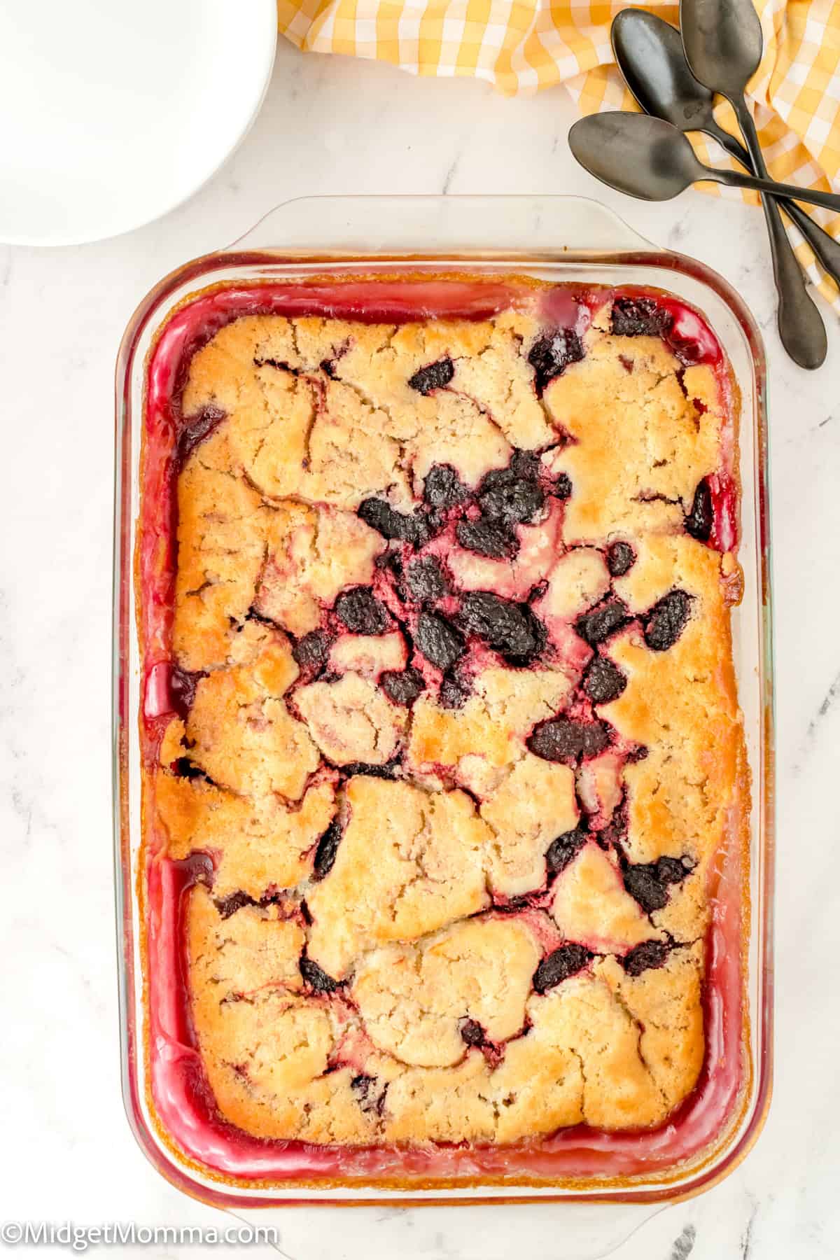 A rectangular berry cobbler in a glass baking dish, with a golden crust and scattered dark berry filling. Spoons and a yellow checkered cloth are beside the dish on a white marble countertop.