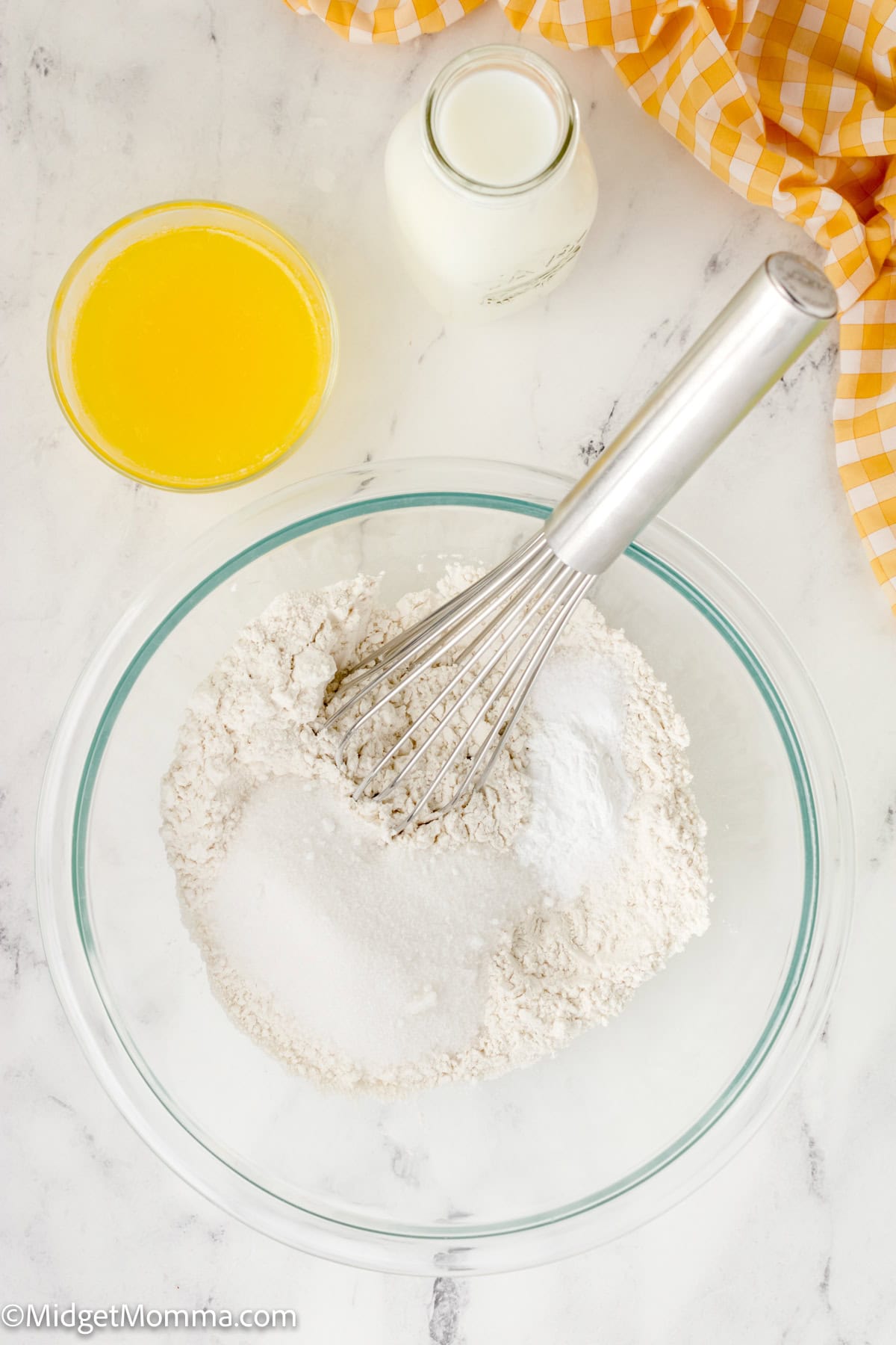 A glass bowl with flour, sugar, and a whisk. Nearby are a glass of orange juice, a small bottle of milk, and a checked cloth on a marble countertop.