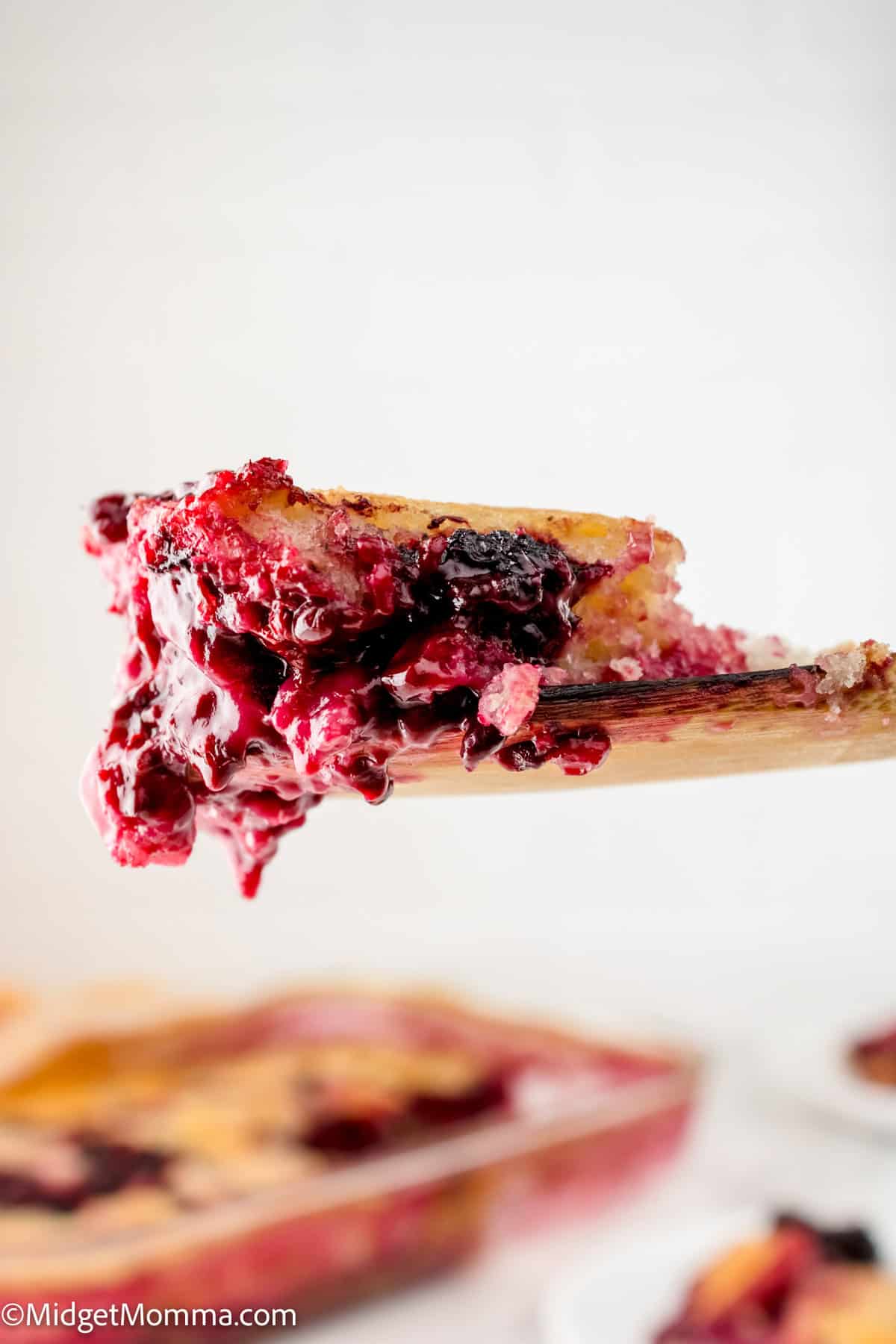 A close-up of a wooden spoon holding a slice of mixed berry cobbler, with a baked dish of cobbler in the background.