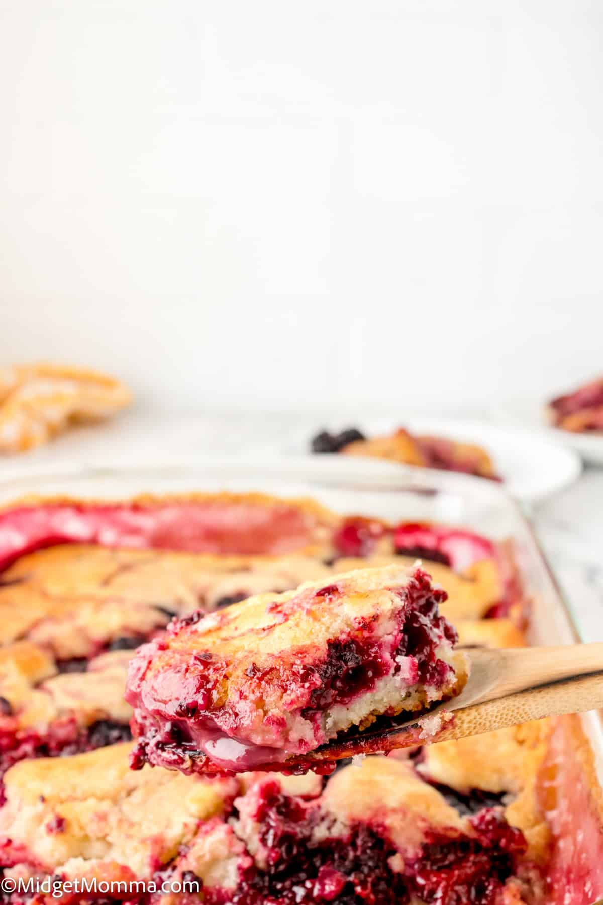 A close-up of a wooden spoon holding a serving of berry cobbler from a larger dish in the background. The cobbler has a golden-brown crust with visible berry filling.