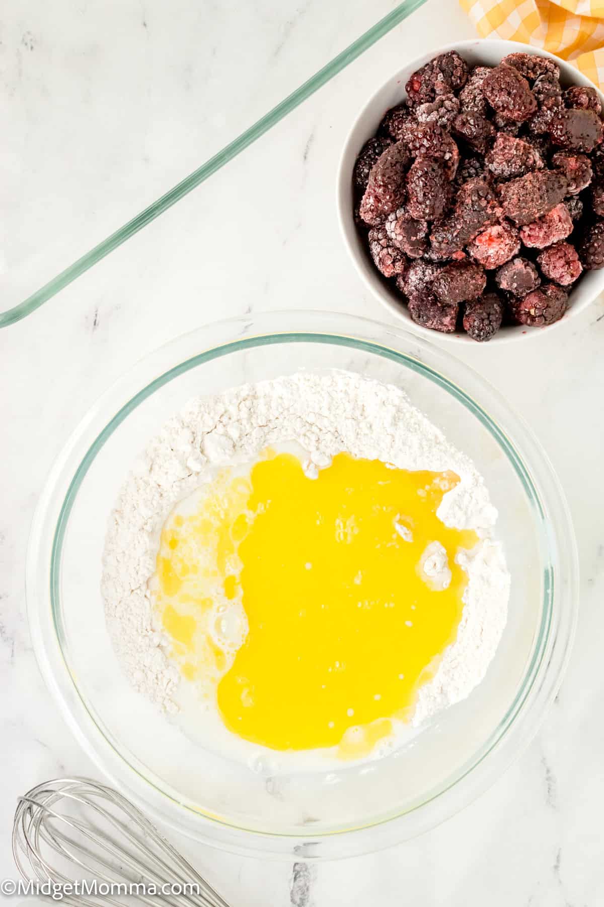 A glass bowl with flour and melted butter mixture next to a bowl of frozen mixed berries on a marble countertop.