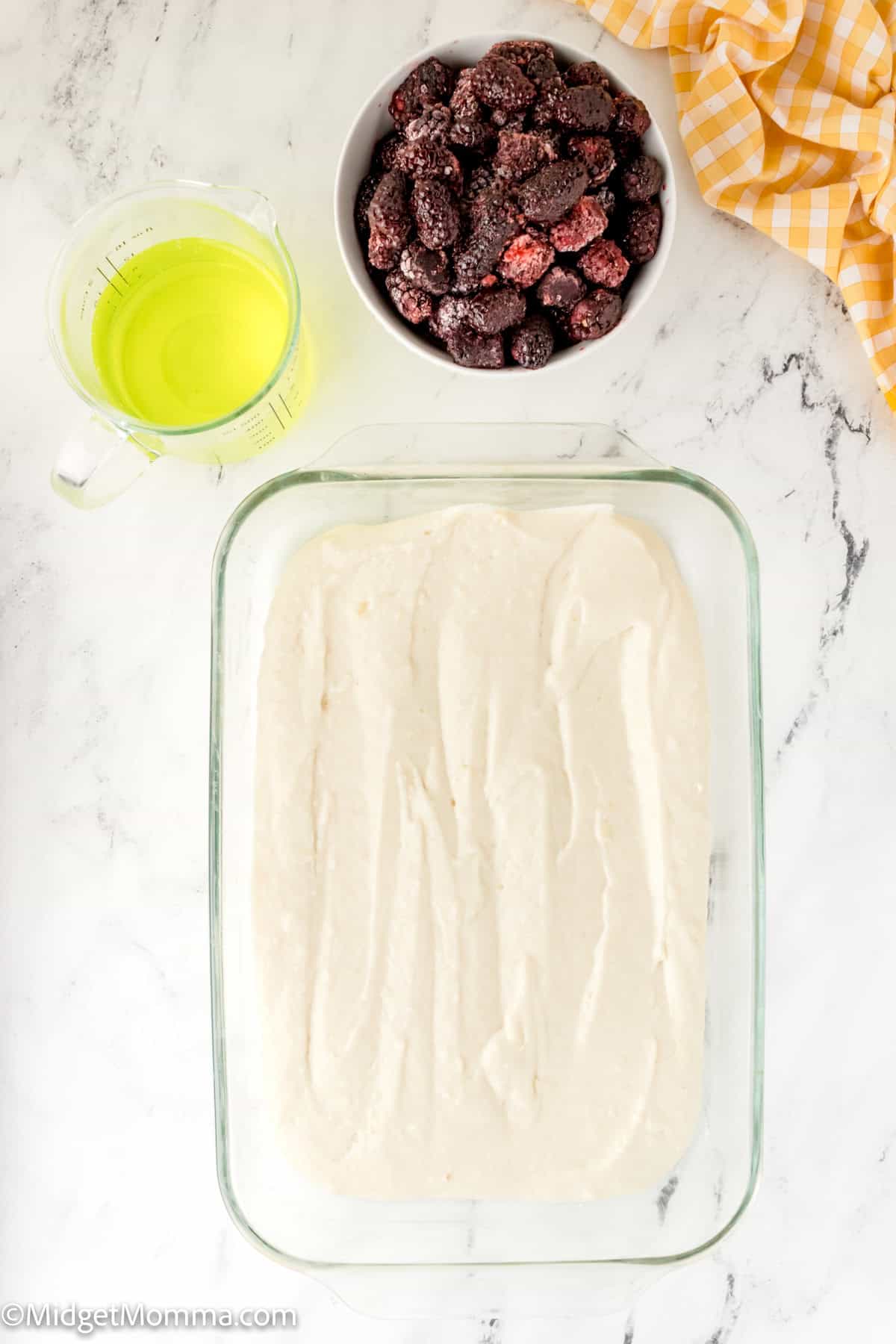 A glass baking dish filled with batter, a bowl of frozen berries, a measuring cup with a yellow liquid, and a yellow checkered cloth on a marble countertop.