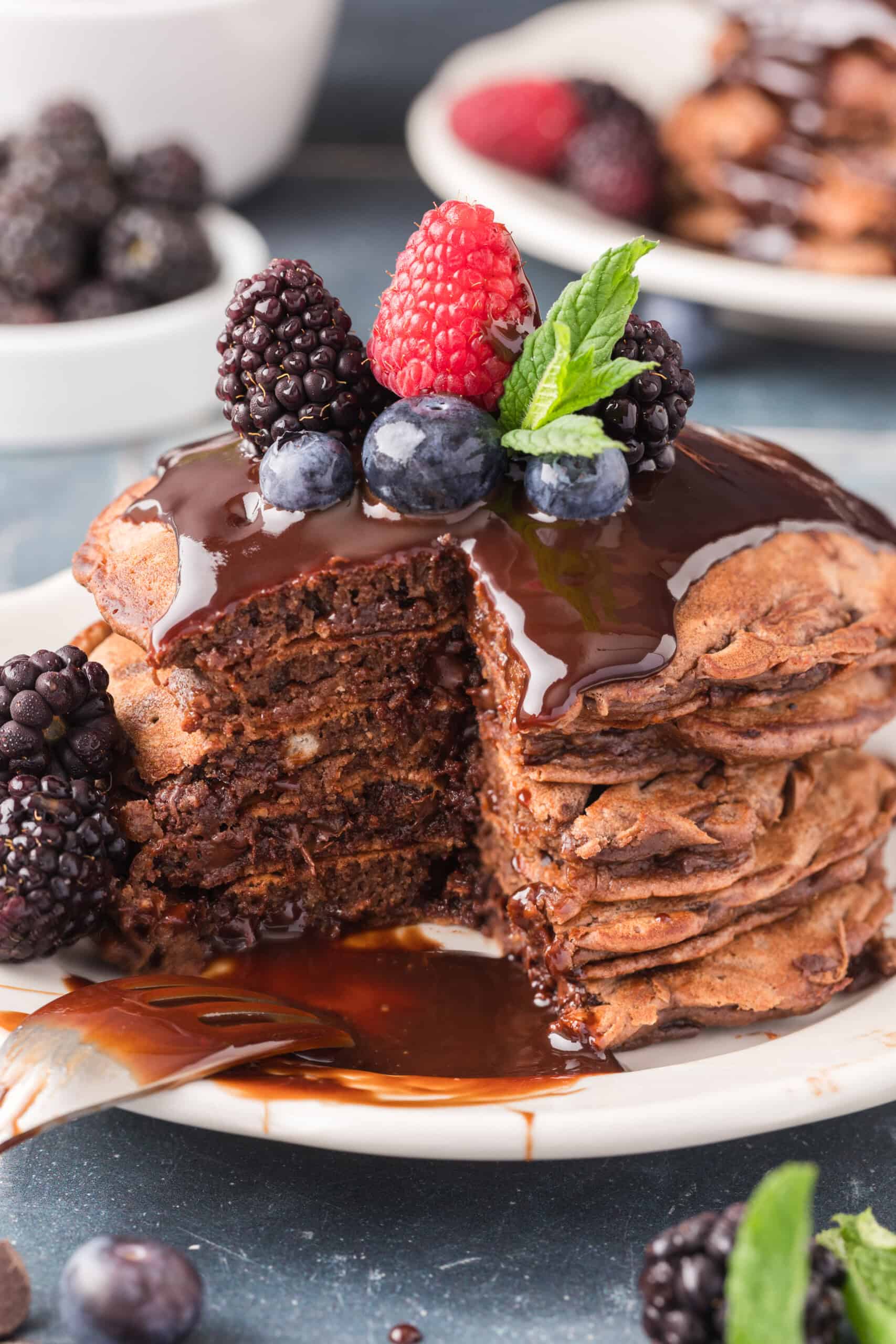 A stack of chocolate pancakes topped with fresh berries and mint, drizzled with chocolate syrup, with a slice cut out on a white plate.