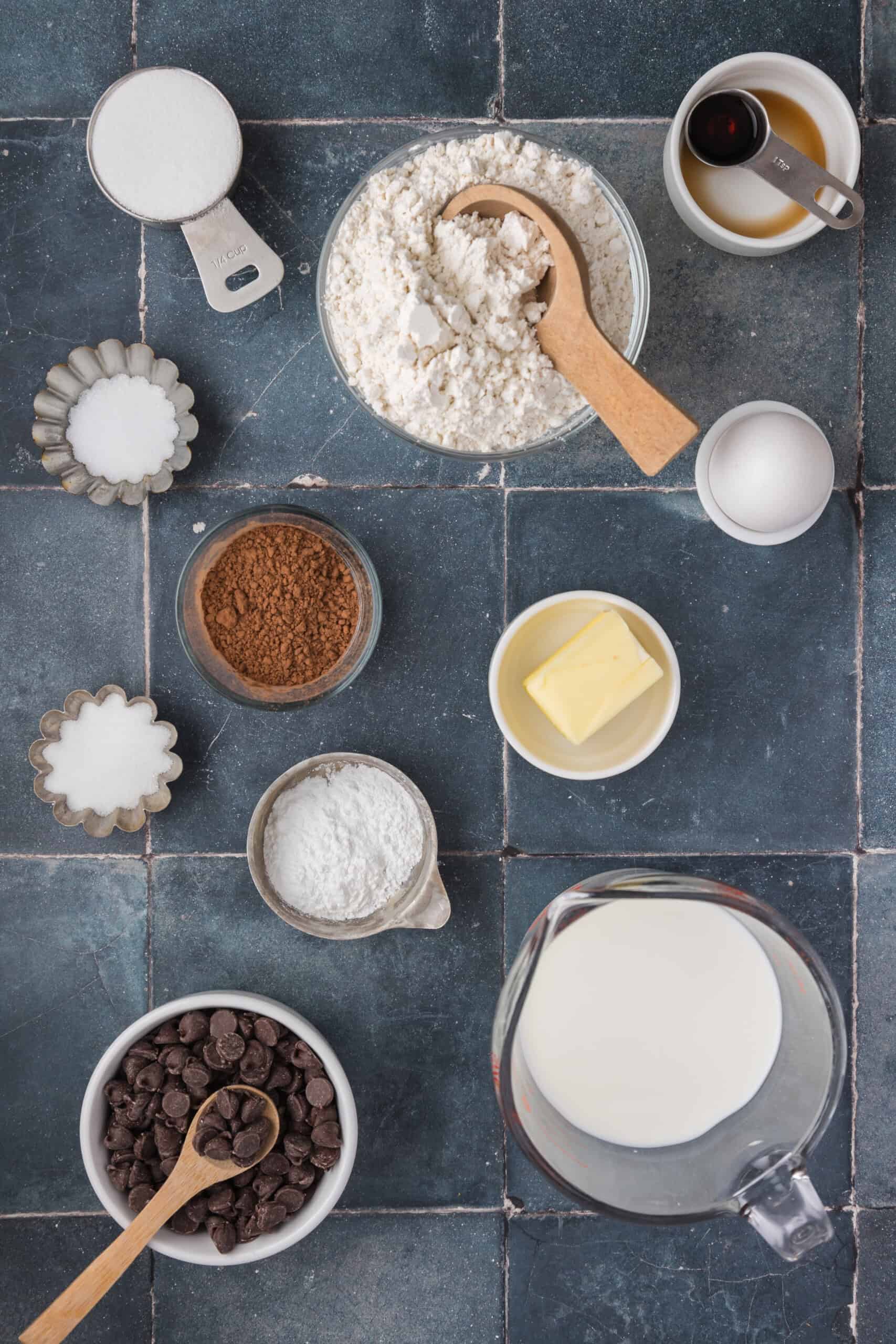 chocolate pancake recipe ingredients. A variety of baking ingredients arranged on a tiled surface, including flour, sugar, butter, cocoa powder, chocolate chips, an egg, milk, and a measuring spoon with vanilla extract.