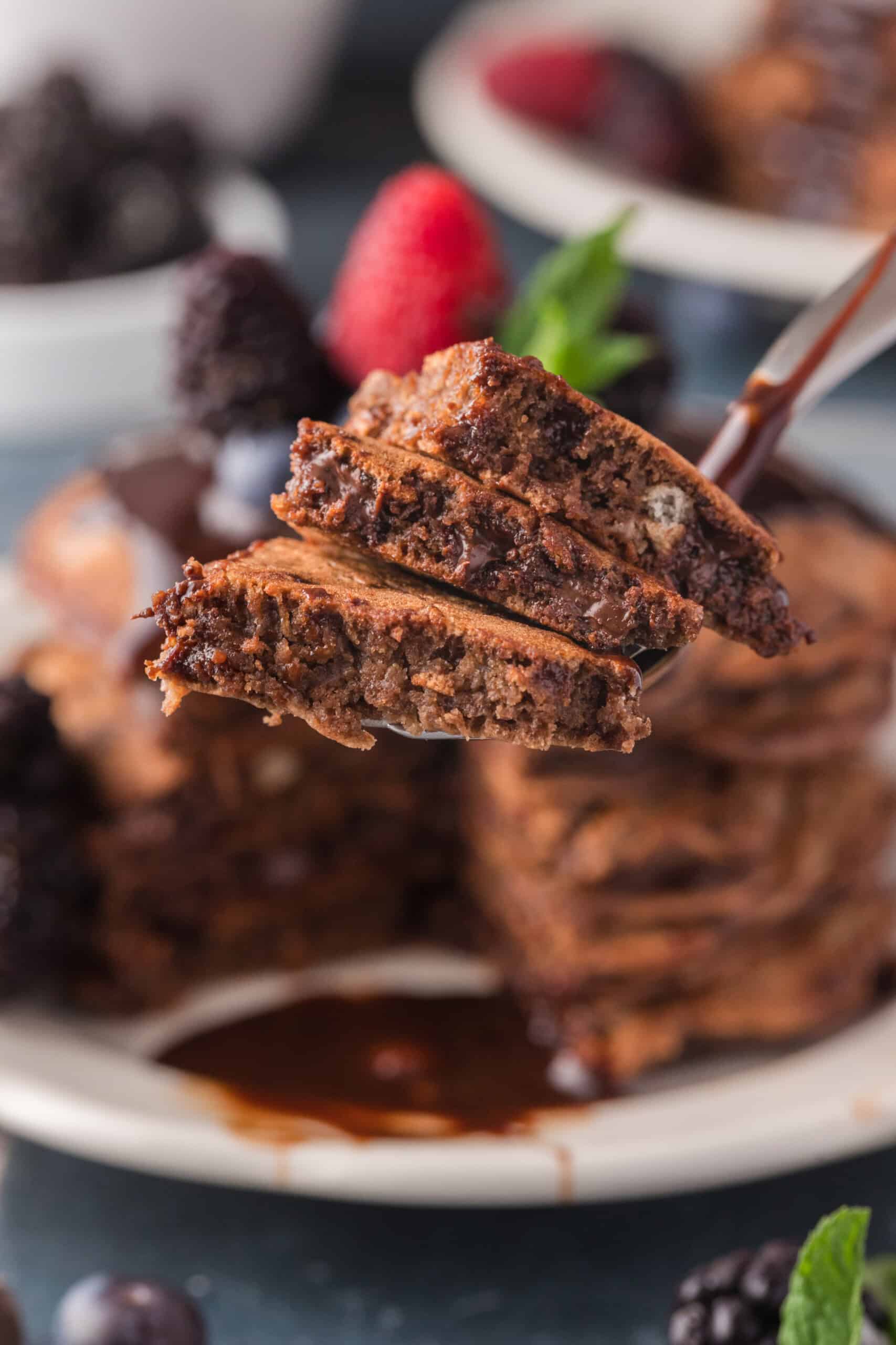 A fork lifts a stack of chocolate pancakes drizzled with syrup from a plate, surrounded by mixed berries.