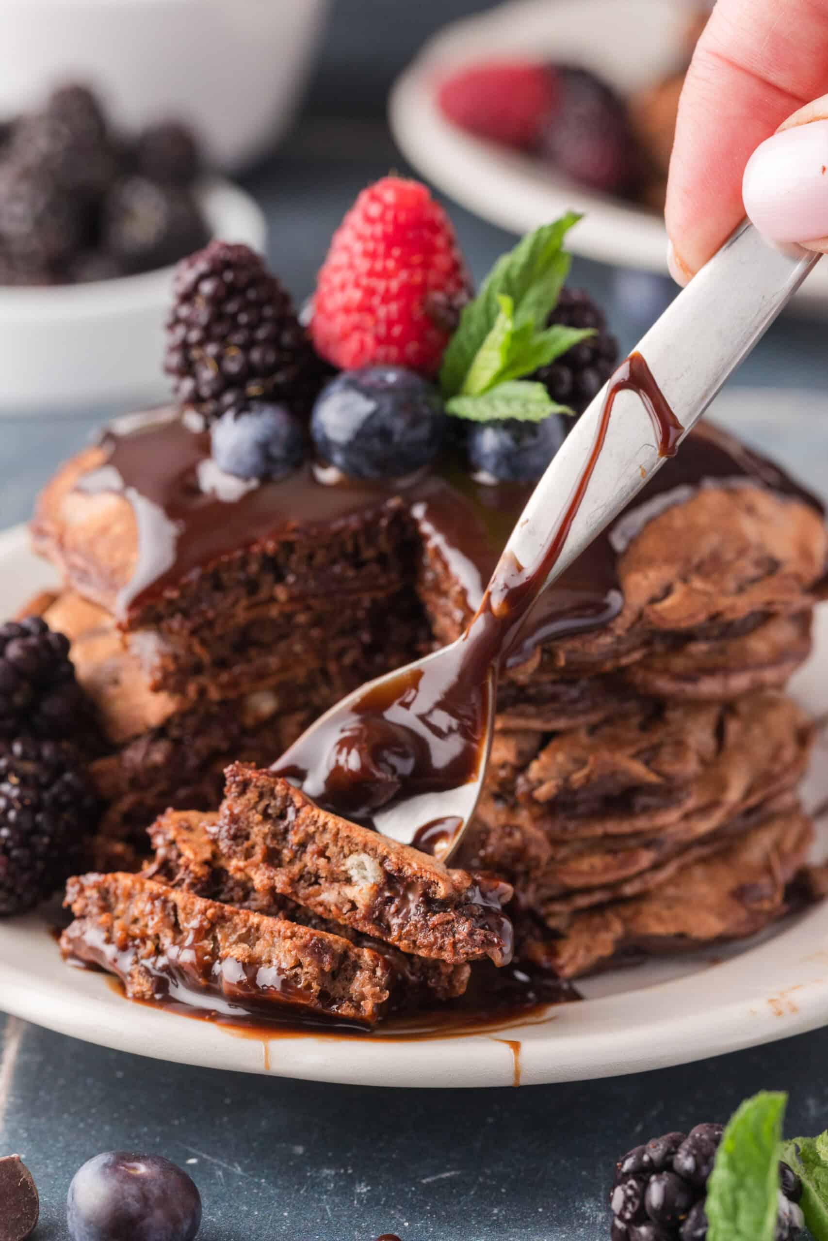 A stack of chocolate pancakes topped with chocolate syrup, blackberries, blueberries, and a raspberry. A fork is holding a piece of the pancakes.