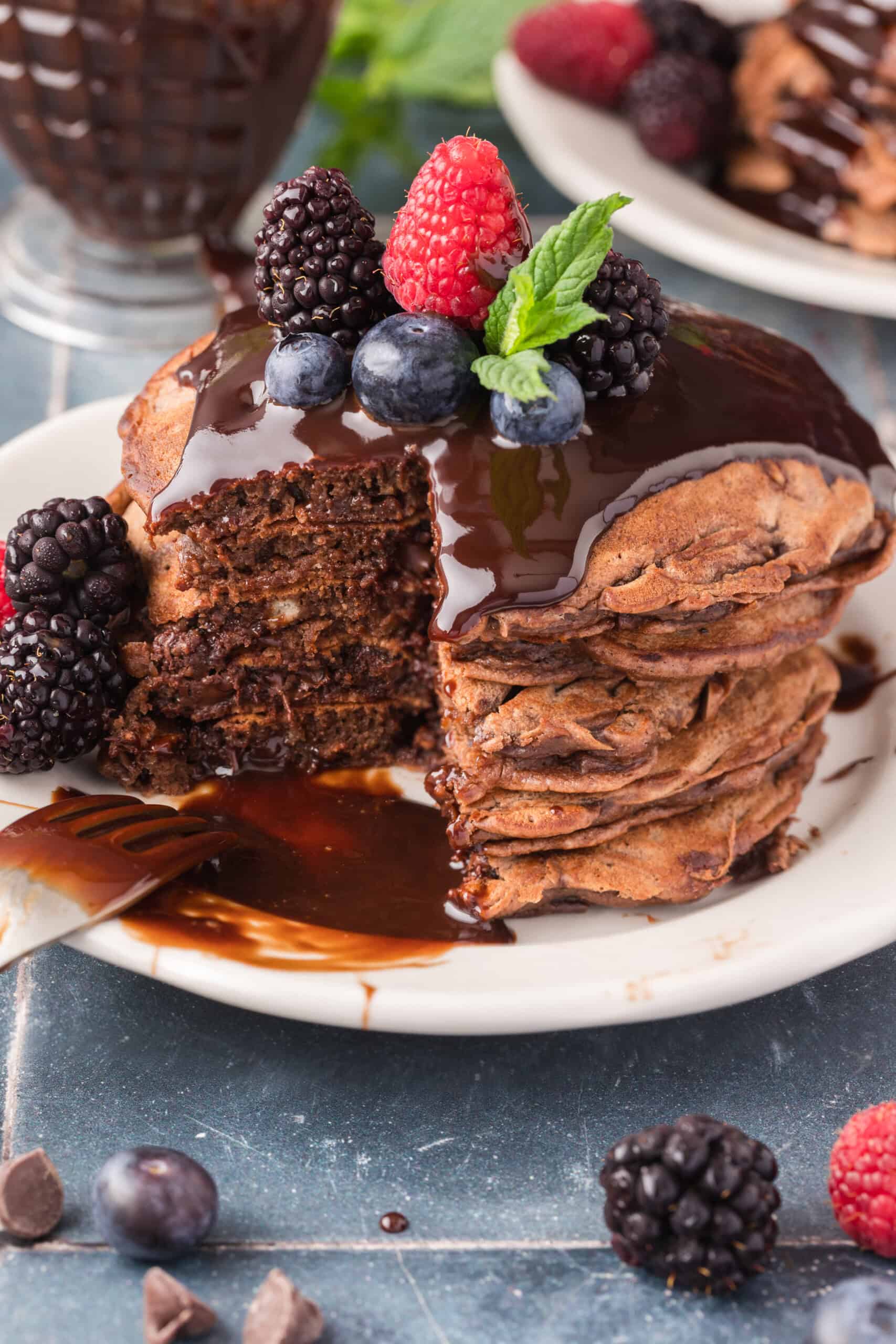 A stack of chocolate pancakes topped with berries and mint leaves is partially sliced. They are drizzled with chocolate syrup. Additional berries are scattered on the table around the plate.