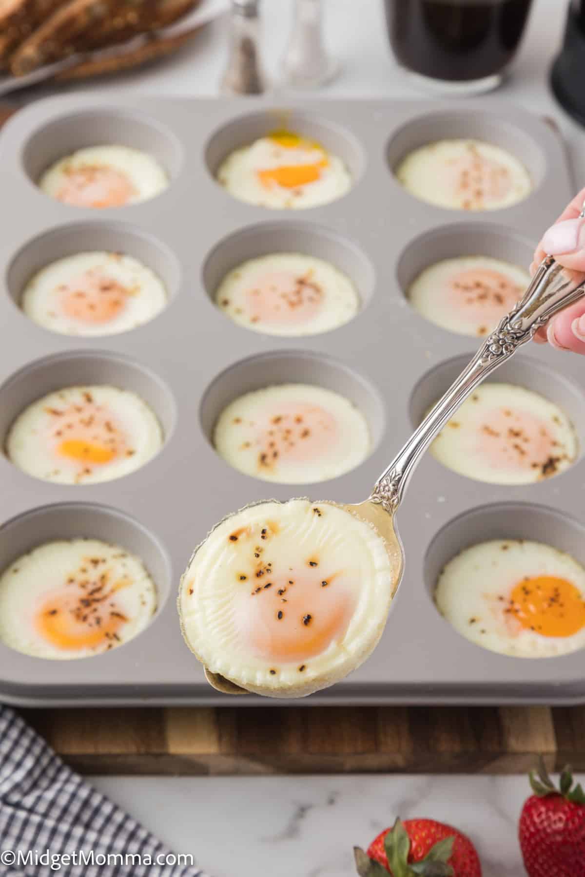 A spoon scooping an egg muffin from a muffin tin with several baked egg muffins, garnished with pepper. Strawberries and a checkered cloth are partially visible.