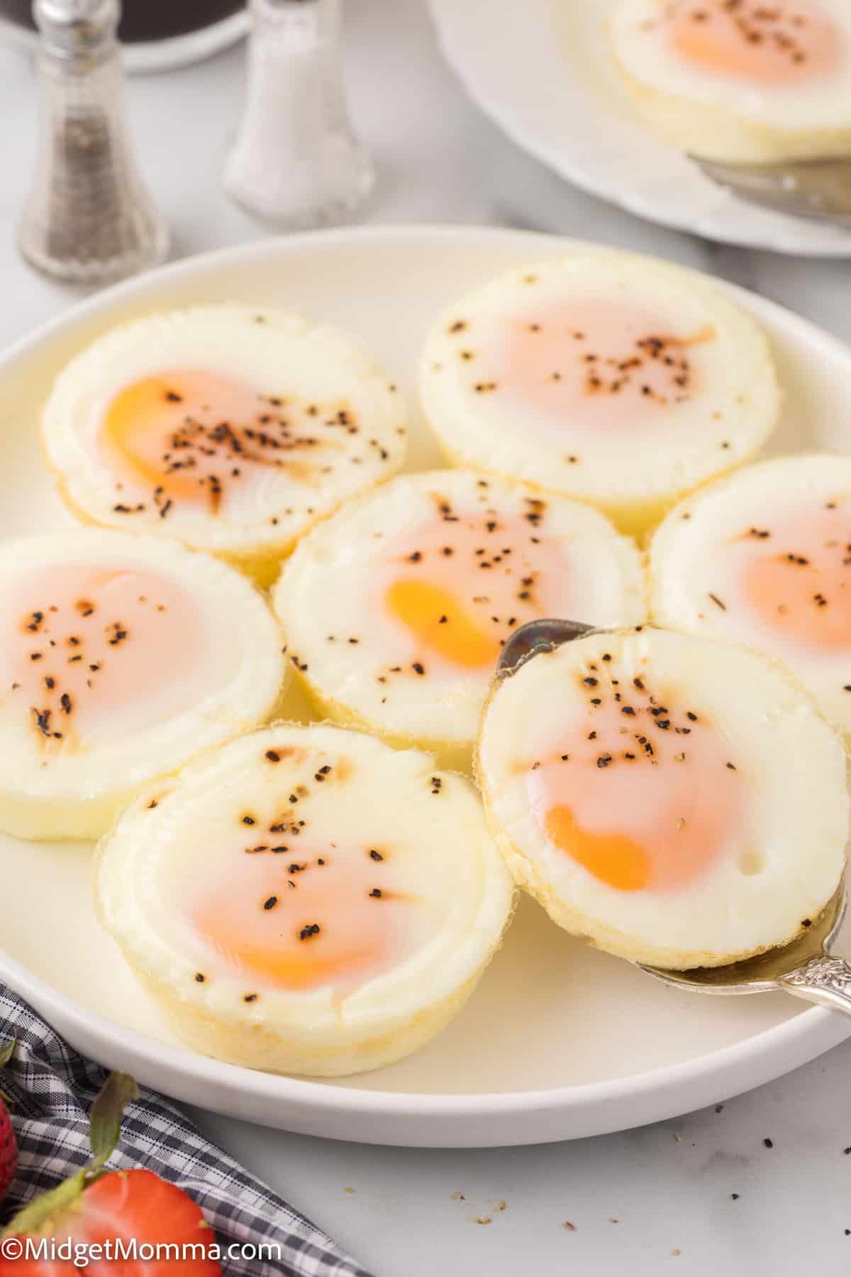 A plate of six muffin tin oven baked eggs with runny yolks, garnished with black pepper. A silver spoon is holding one egg. A strawberry and pepper shakers are seen at the edges.