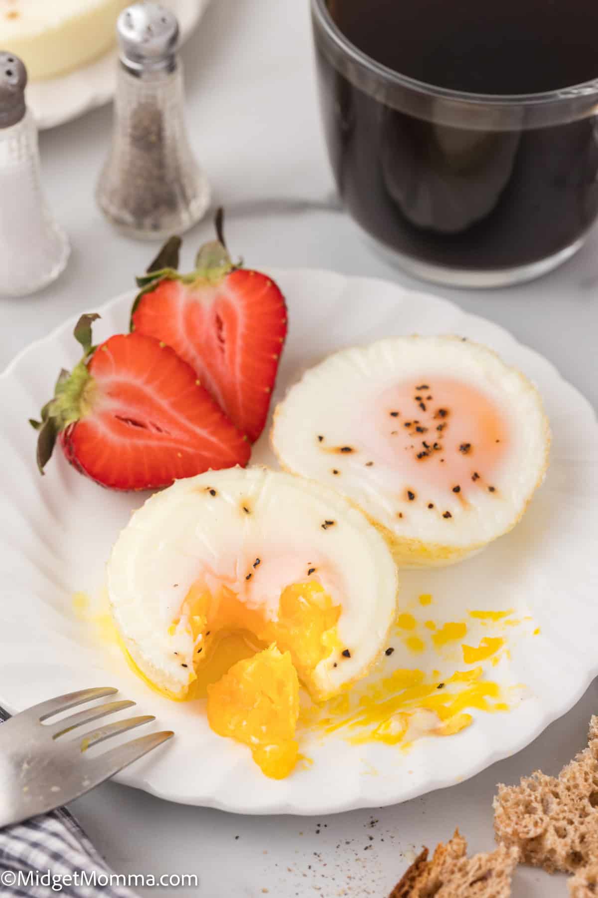 A white plate with two halved soft-boiled eggs sprinkled with pepper, two slices of strawberry, a fork, and a cup of black coffee in the background.