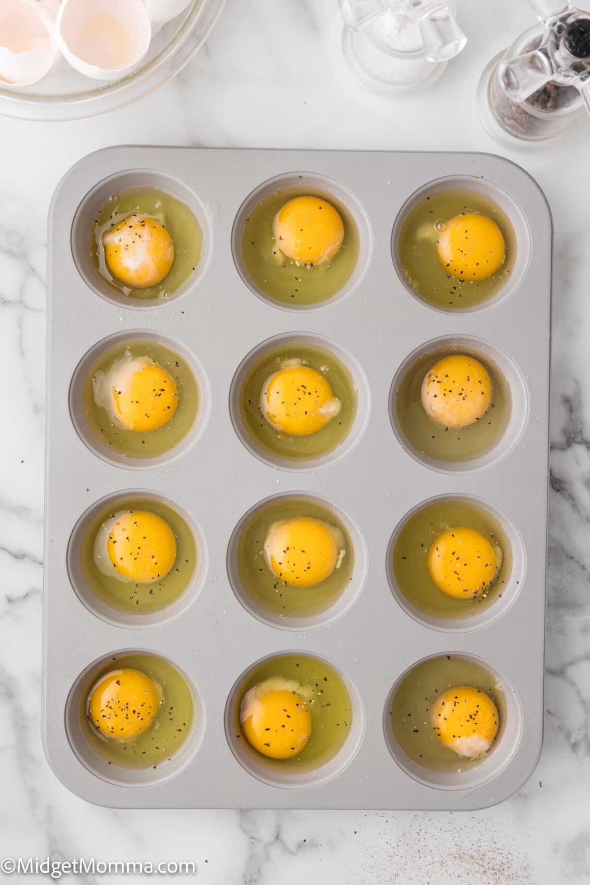 A muffin tin filled with twelve raw eggs cracked into each slot, seasoned with salt and pepper, on a marble countertop.