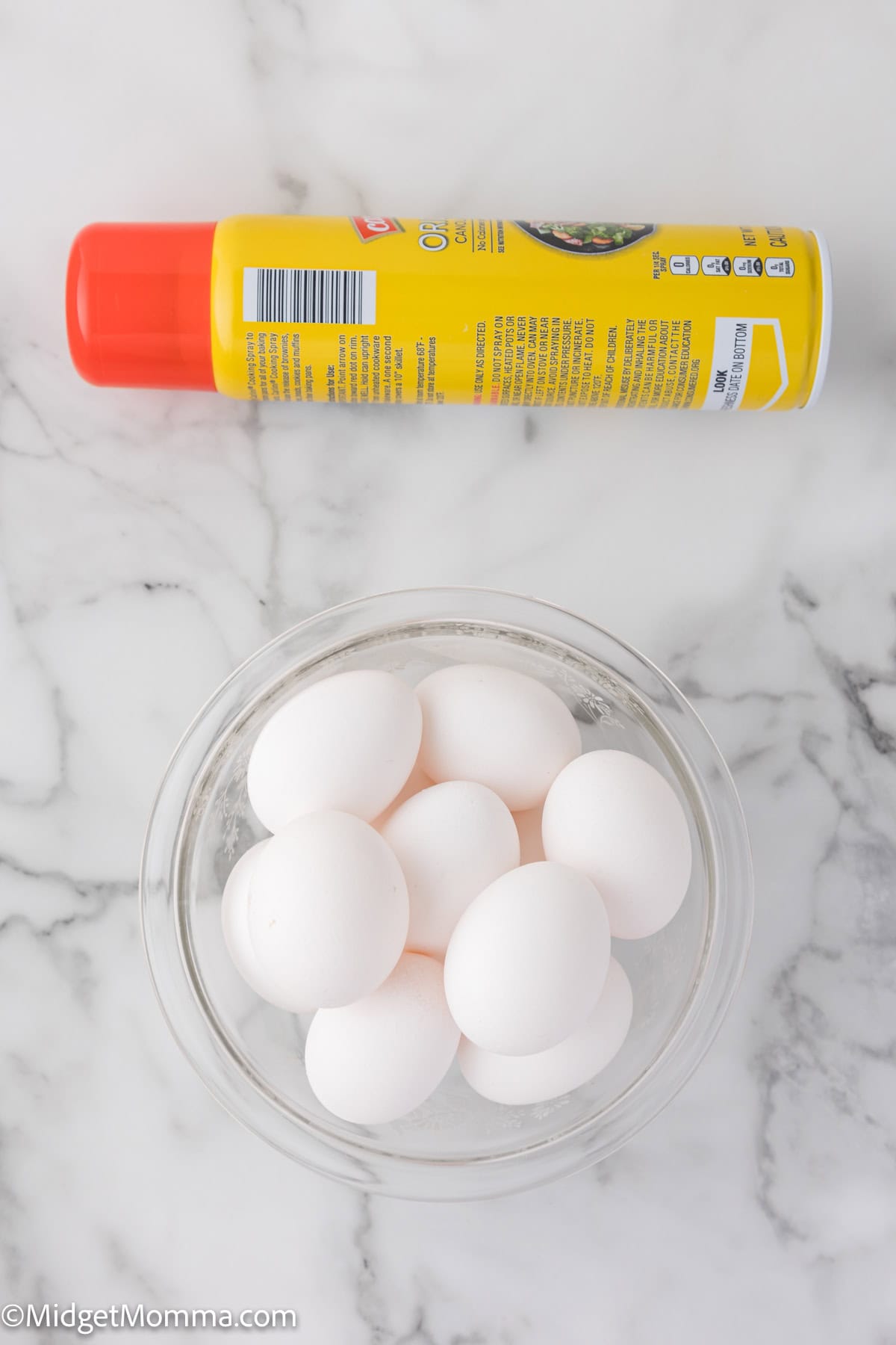 A bowl of white eggs sits on a marble countertop next to a can of cooking spray with a red cap.