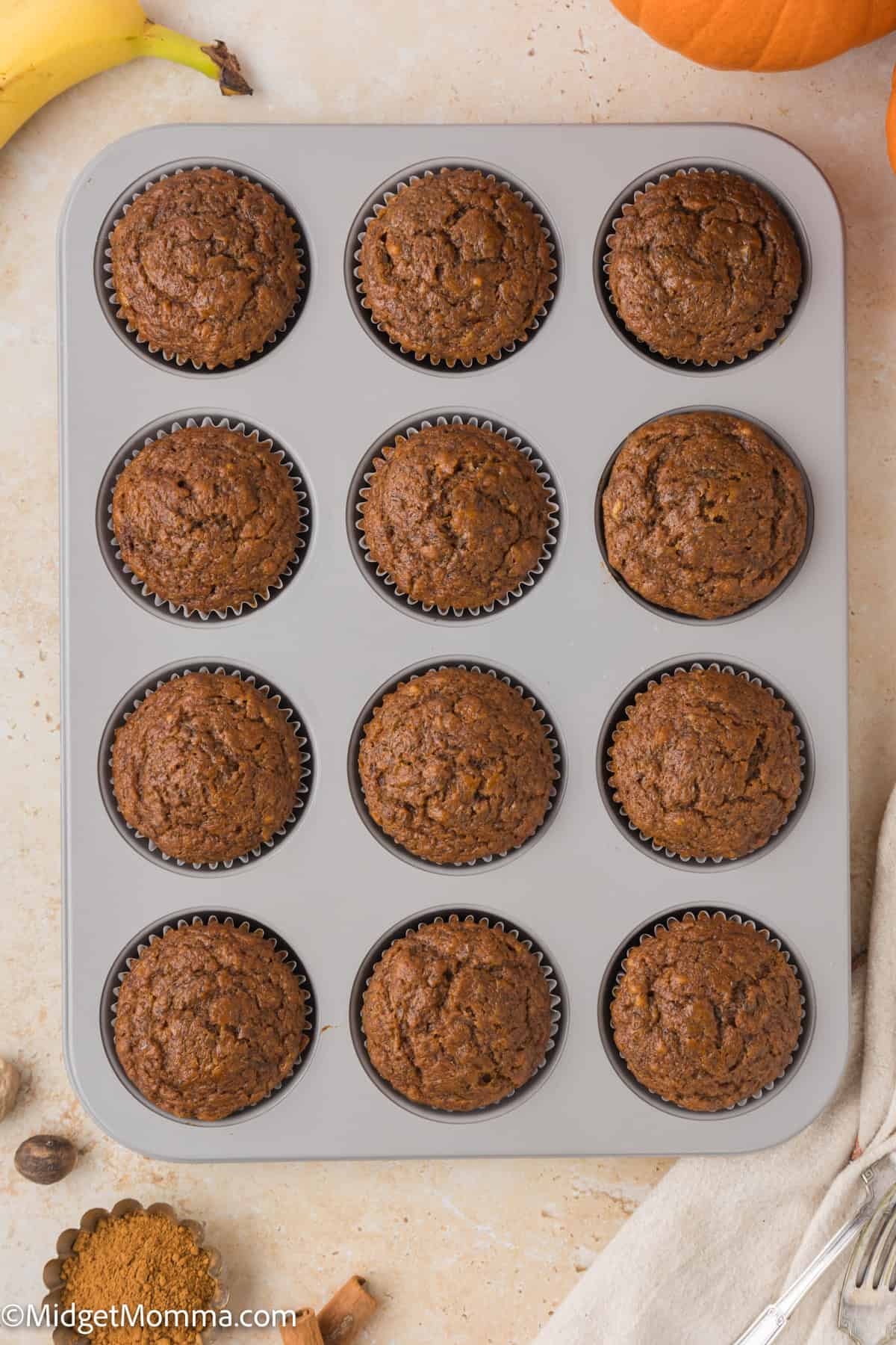 A gray muffin tin holding twelve baked chocolate muffins on a light-colored surface with a few scattered baking ingredients and utensils.