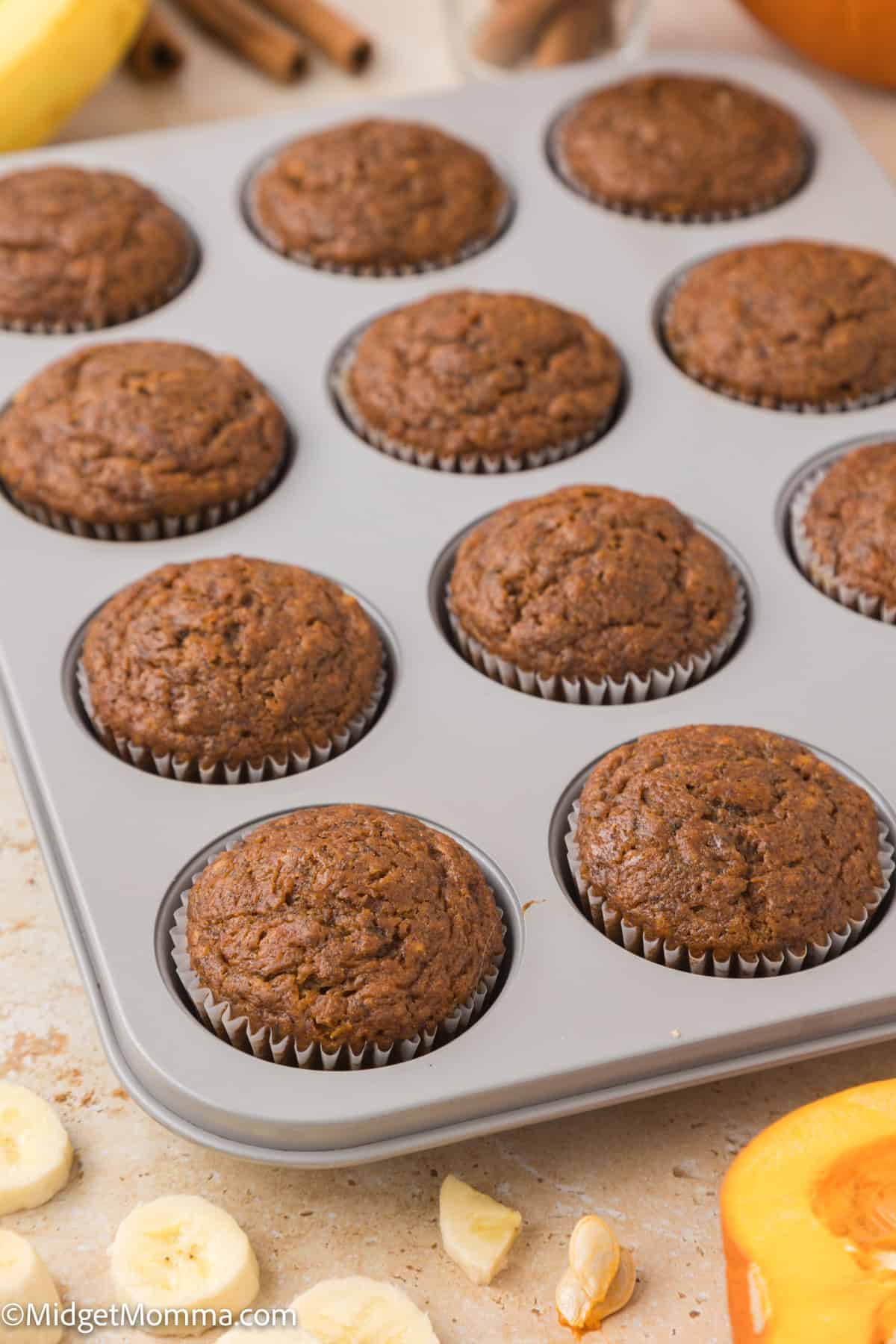 A muffin tray filled with twelve baked muffins. Slices of bananas and pieces of nuts are scattered around on a beige surface.