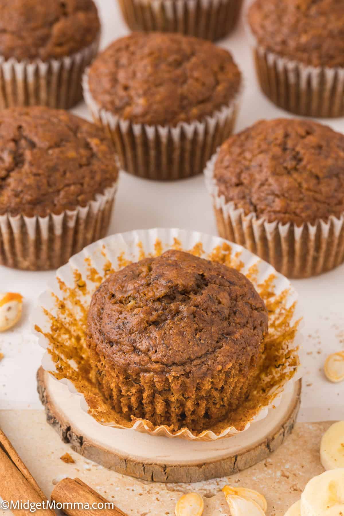 A group of muffins with one partially unwrapped muffin in the foreground, placed on a wooden coaster. 