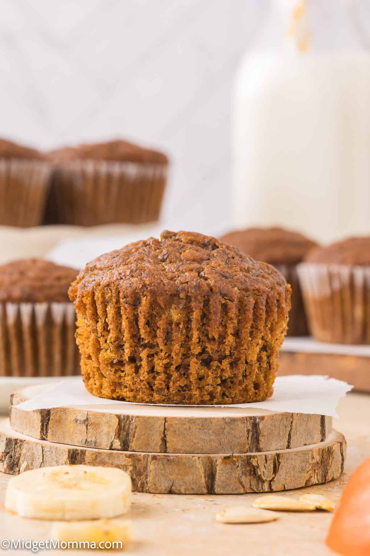 A banana muffin is displayed on a wooden slice, with more muffins and a milk bottle blurred in the background.