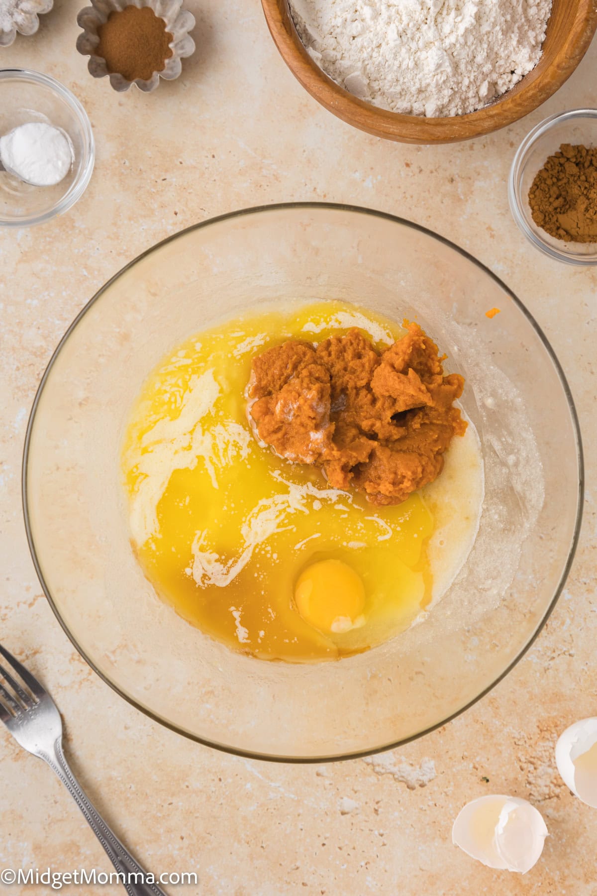 A mixing bowl containing wet baking ingredients, including an egg, pumpkin puree, and melted butter. Surrounding the bowl are various dry ingredients like flour, spices, and an empty cracked eggshell.