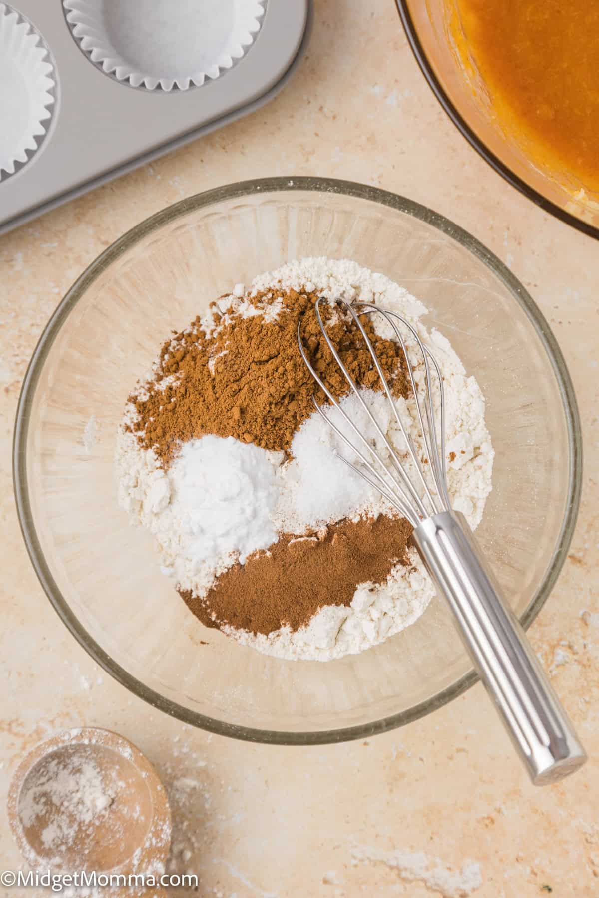 A glass bowl contains flour, brown sugar, baking powder, and spices with a whisk. Nearby are a muffin tin with liners, a glass bowl.