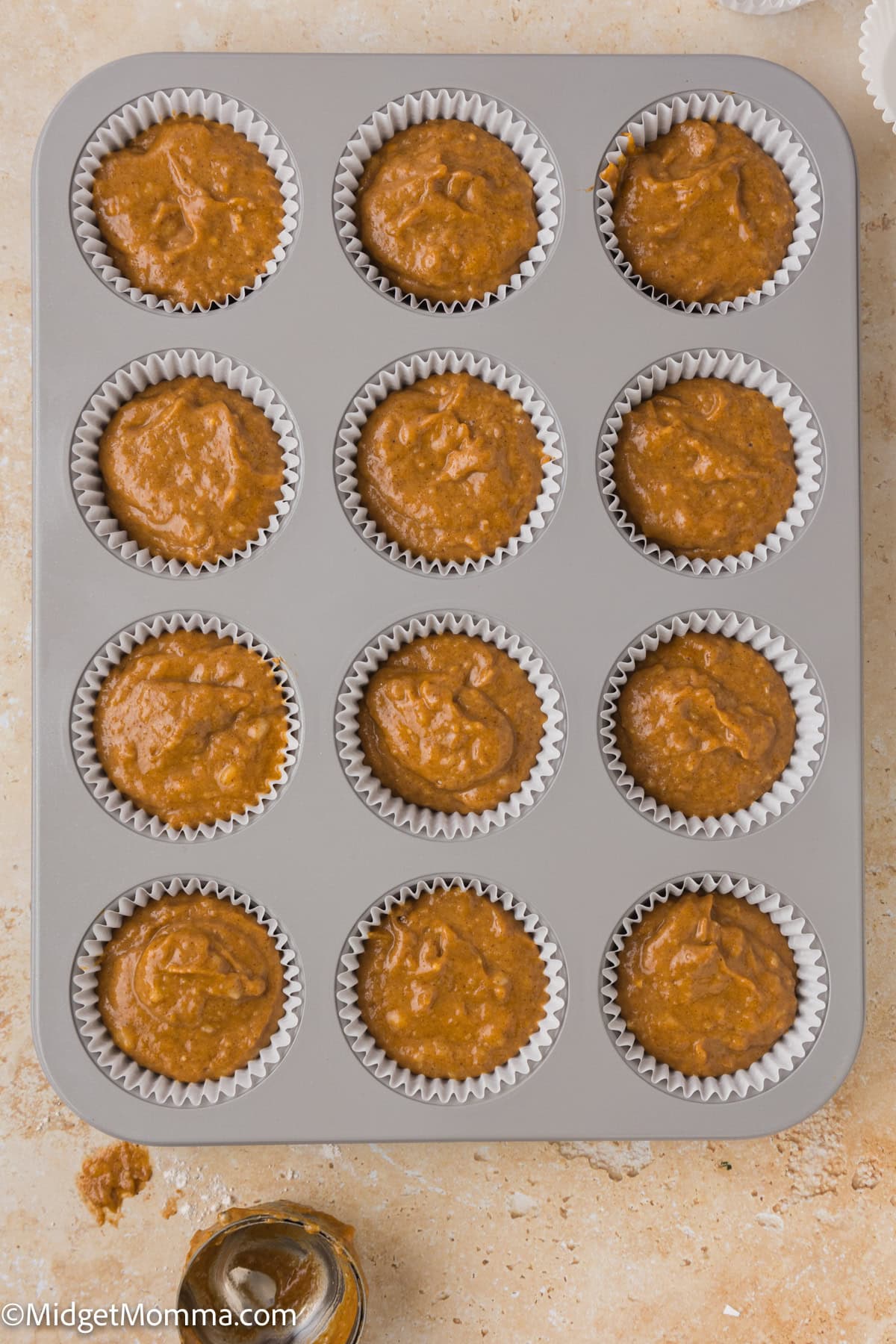 A muffin tin containing twelve unbaked muffins in white paper liners, placed on a beige countertop.