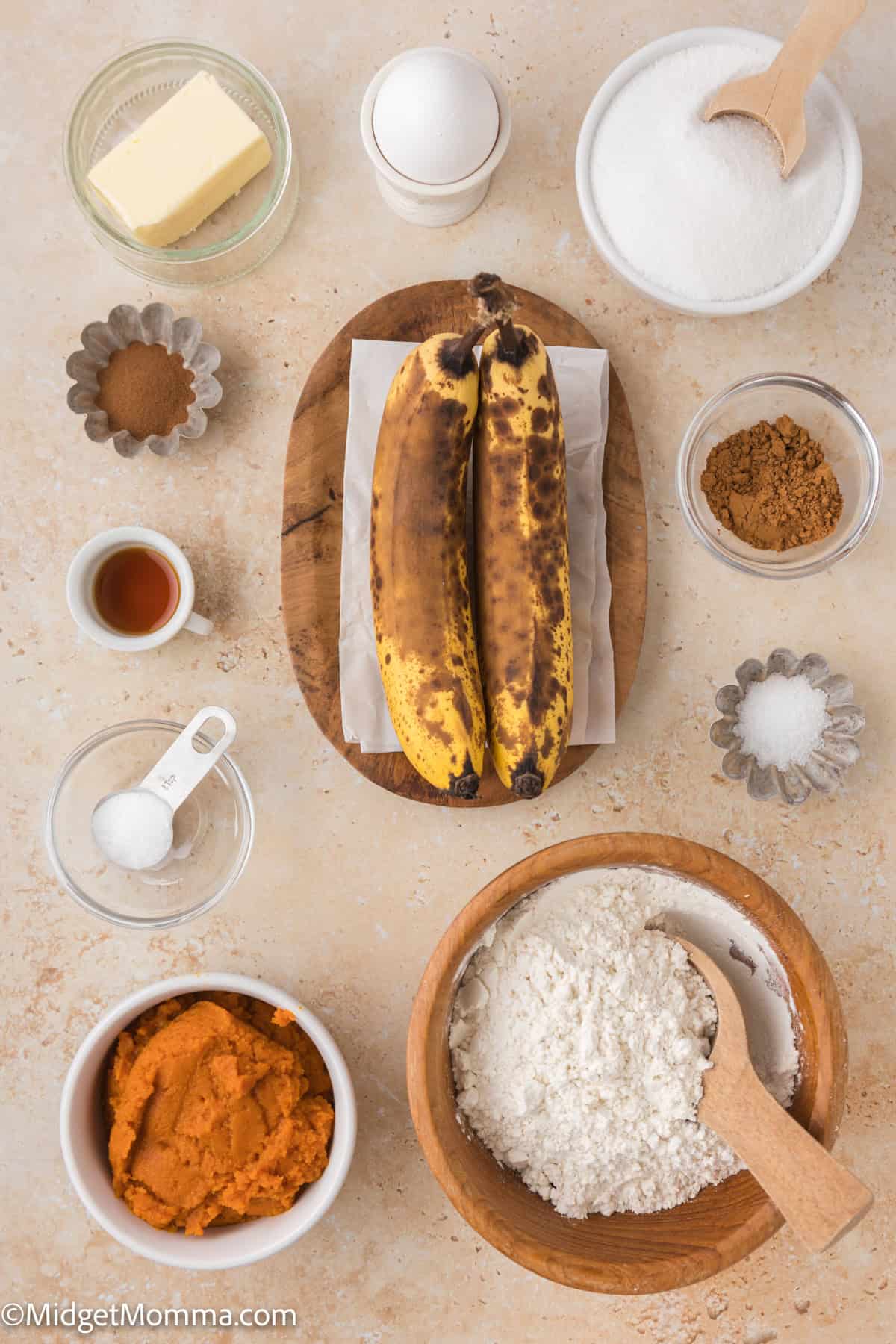 Ingredients for baking banana bread arranged on a countertop, including ripe bananas, flour, sugar, butter, egg, vanilla extract, cinnamon, salt, baking soda, and pumpkin puree.