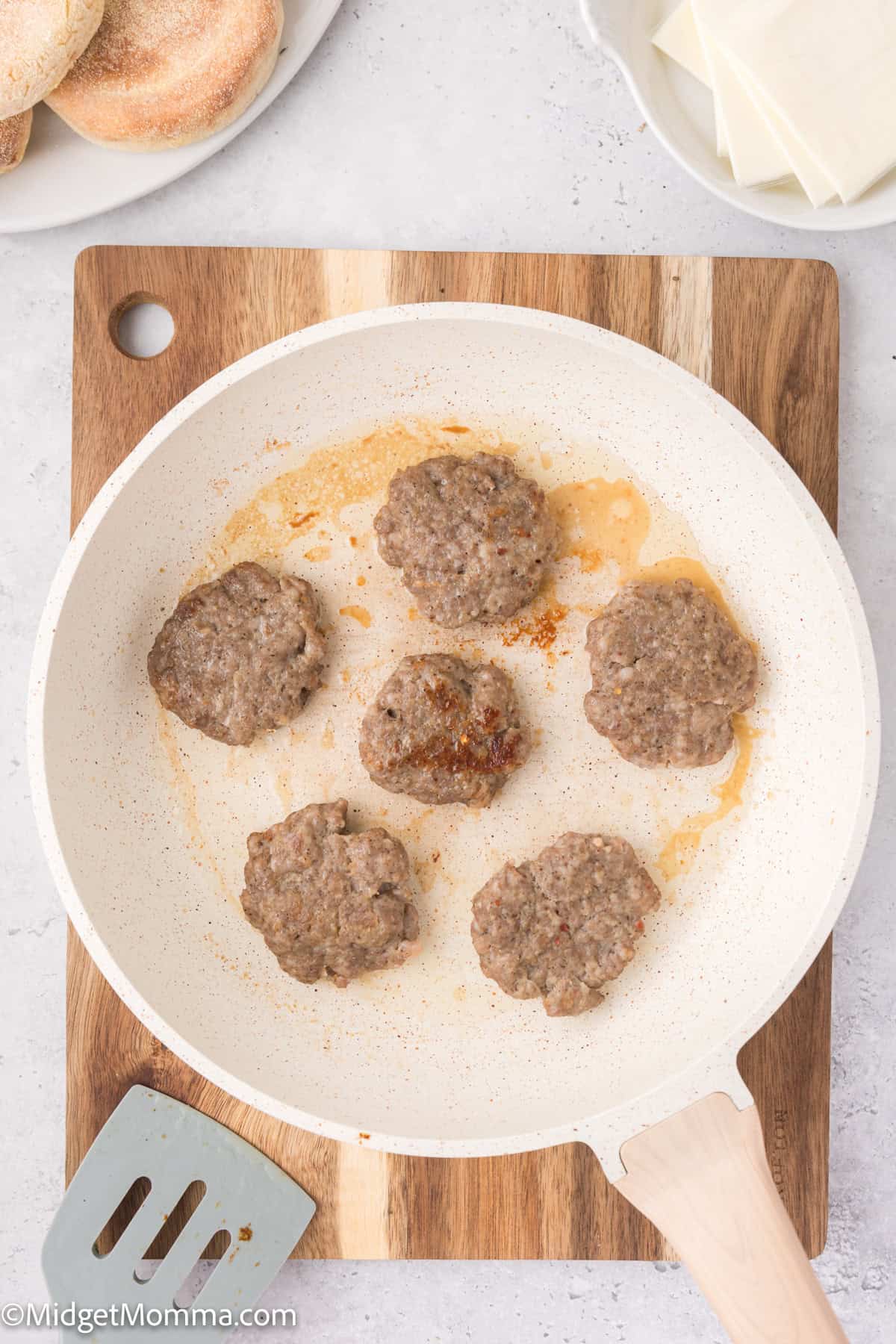 A white skillet with six cooking sausage patties is placed on a wooden board. A metal spatula is on the side, and slices of cheese and a few bread rolls are visible near the skillet.