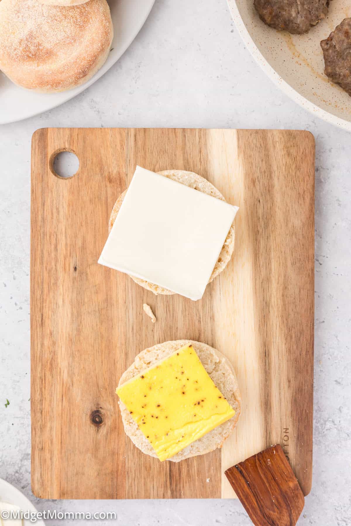 Two English muffin halves on a wooden cutting board, one topped with a slice of white cheese and the other with a slice of yellow cheese. A plate with a bread roll and a pan with meat patties are visible nearby.