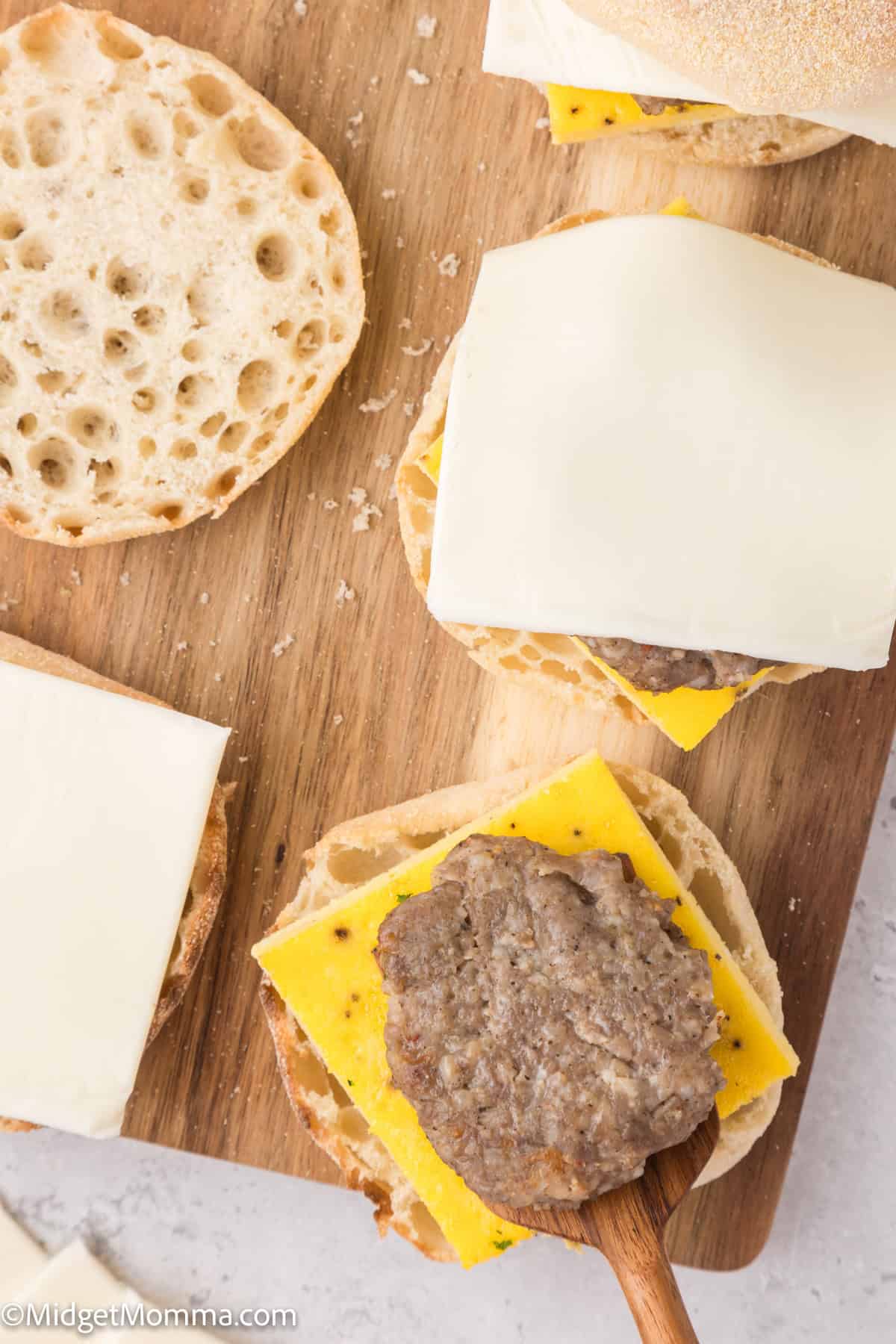 An open English muffin with cheese and sausage patty on a wooden board next to another muffin half with a cheese slice.