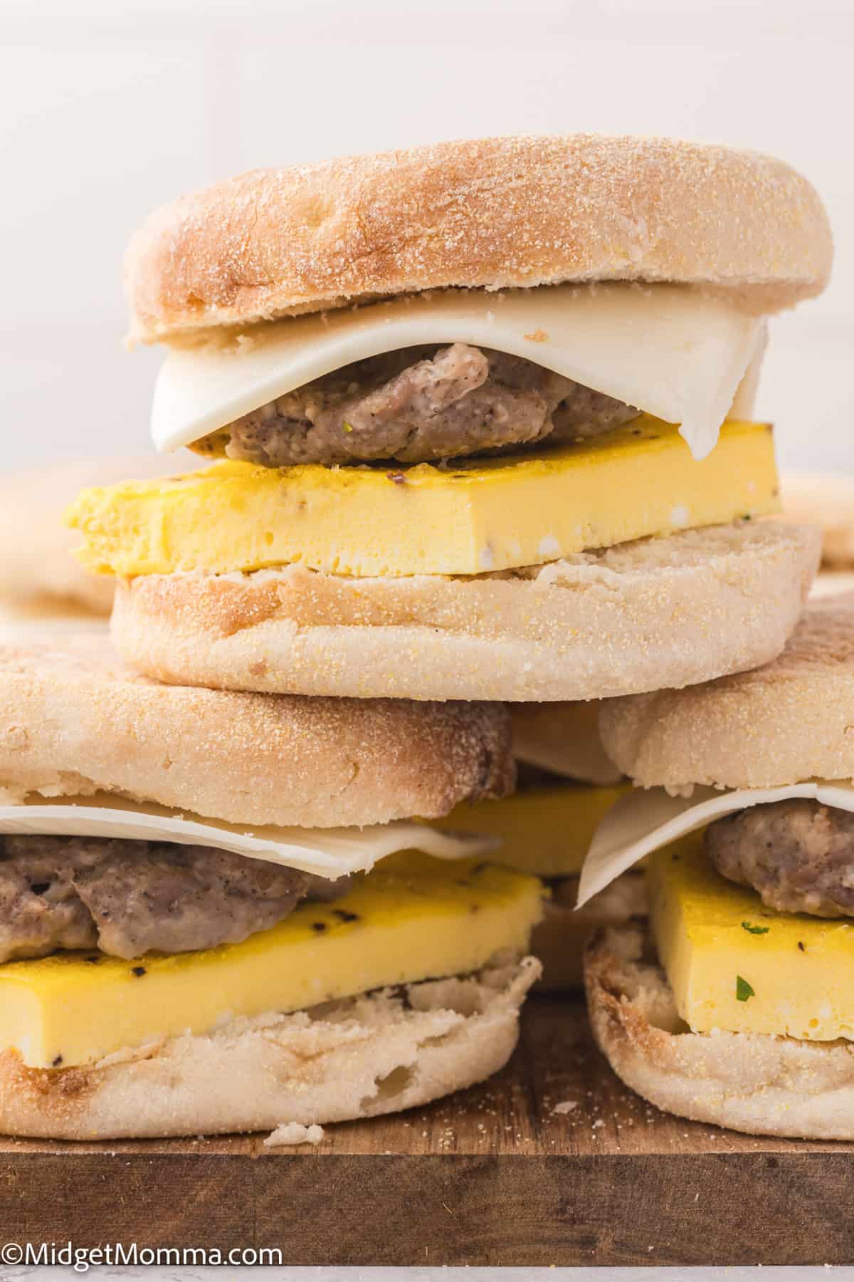 A stack of homemade breakfast sandwiches with English muffins, egg, sausage patty, and cheese is displayed on a wooden surface.