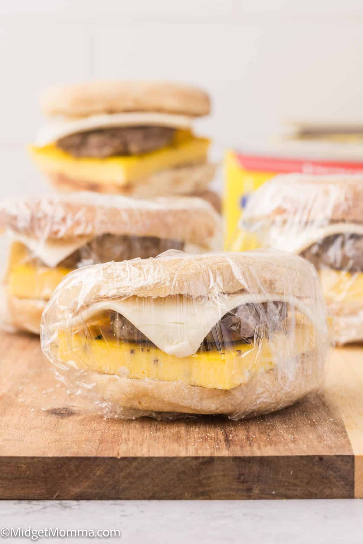 Breakfast sandwiches with eggs, cheese, and sausage wrapped in plastic wrap on a wooden surface.