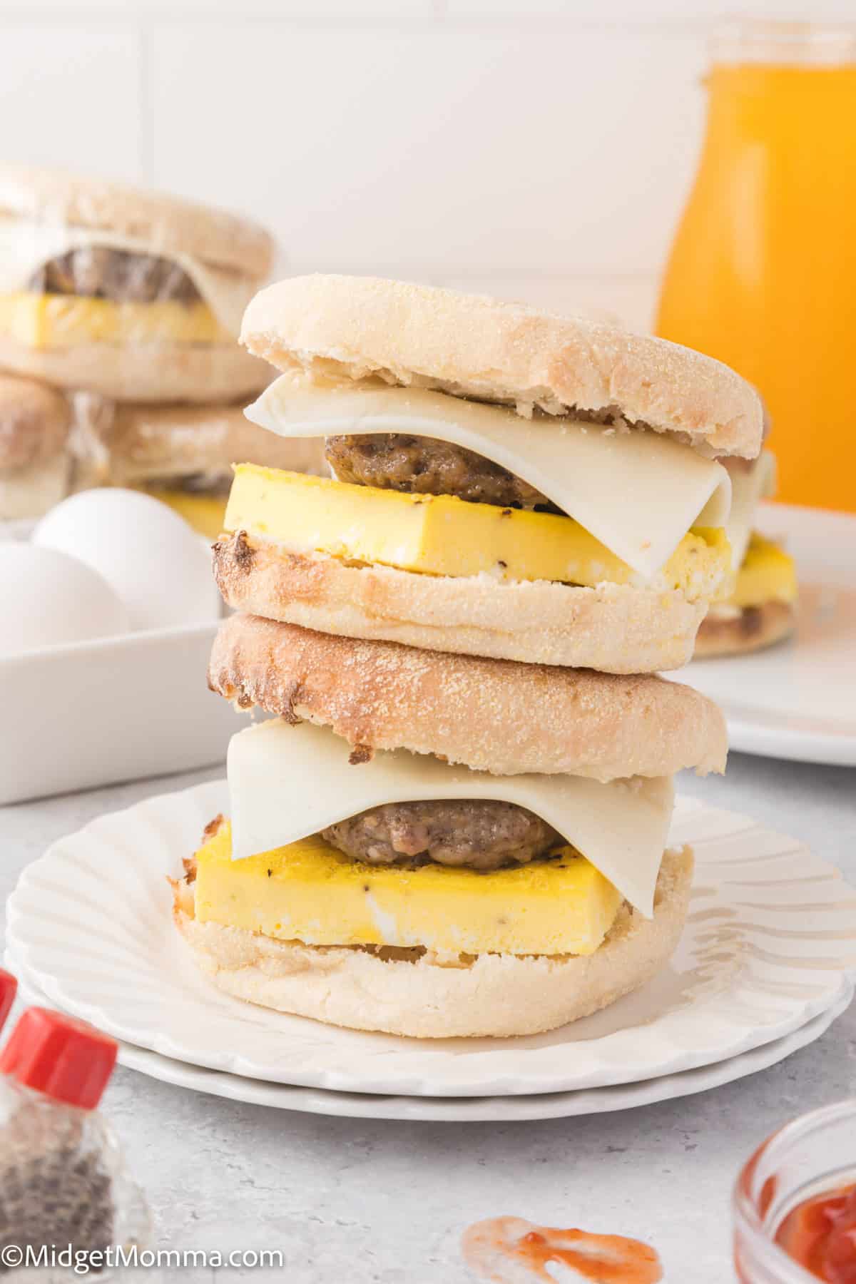 Two breakfast sandwiches with sausage, egg, and cheese are stacked on a white plate. A glass of orange juice and other similar sandwiches are visible in the background.