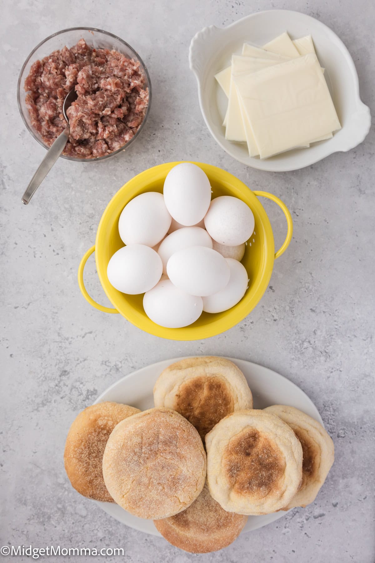 A top-down view of English muffins, a bowl of raw eggs, a dish of cheese slices, and a bowl of sausage meat on a light gray surface.