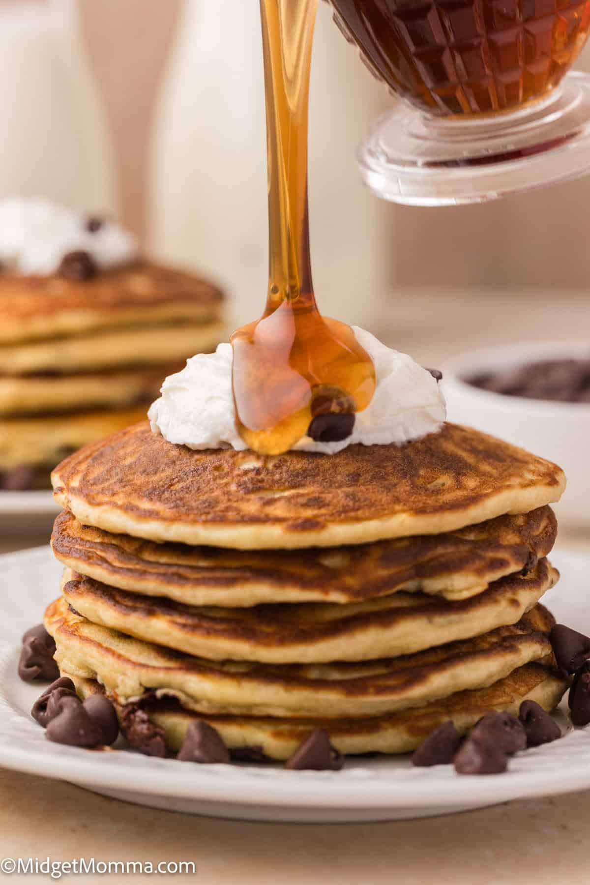 A stack of pancakes topped with whipped cream and chocolate chips is being drizzled with syrup from a container.