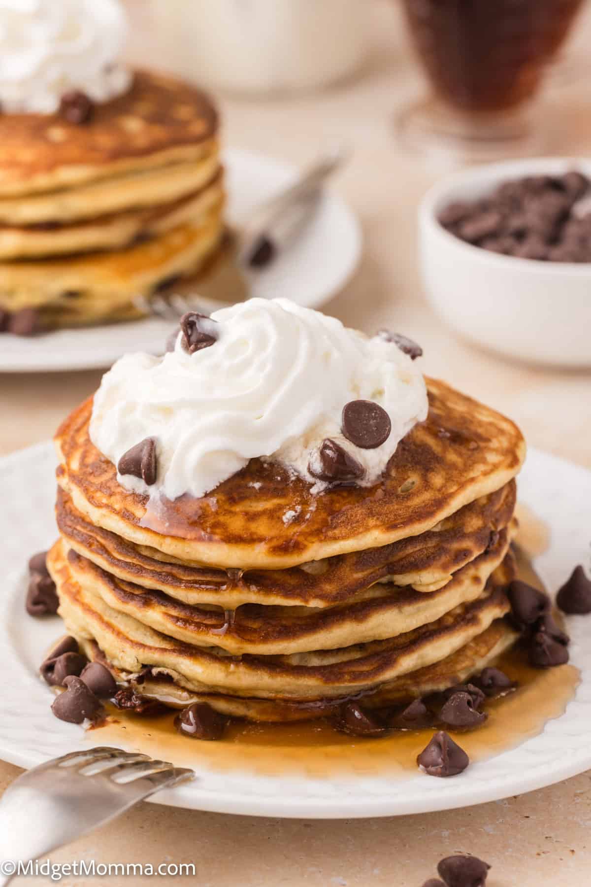 A stack of pancakes topped with whipped cream, chocolate chips, and syrup on a white plate with additional chocolate chips and syrup in the background.