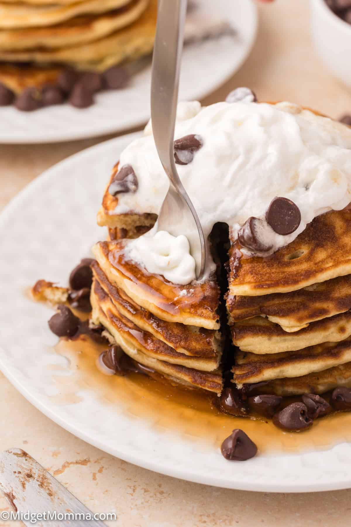 A stack of pancakes topped with whipped cream and chocolate chips on a white plate, with syrup dripping, while a fork slices through the stack.