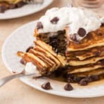 A stack of pancakes topped with whipped cream and chocolate chips is partially eaten on a white plate. A fork is placed on the plate with a piece of the pancake stack.