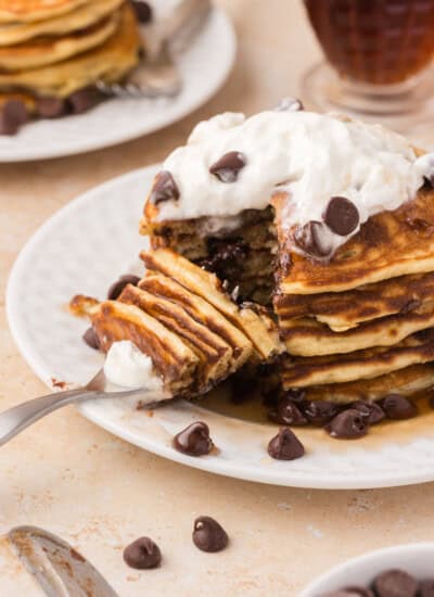 A stack of pancakes topped with whipped cream and chocolate chips is partially eaten on a white plate. A fork is placed on the plate with a piece of the pancake stack.