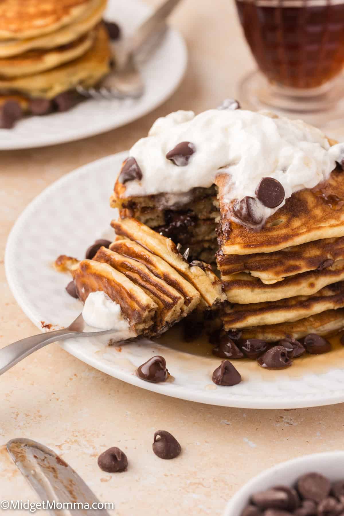 A stack of pancakes topped with whipped cream and chocolate chips is partially eaten on a white plate. A fork is placed on the plate with a piece of the pancake stack.