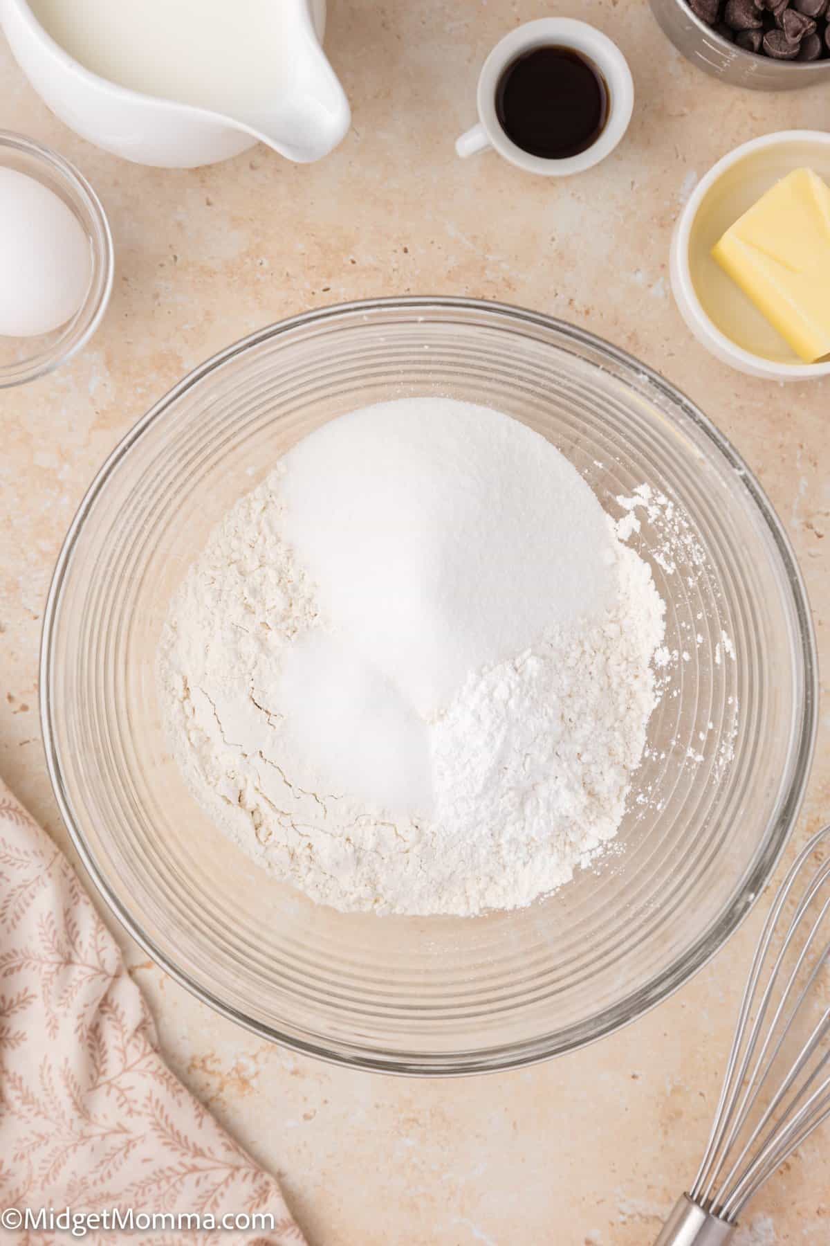 A glass bowl with flour and other dry pancake recipe ingredients, surrounded by a whisk, egg, butter, vanilla, and milk on a countertop.