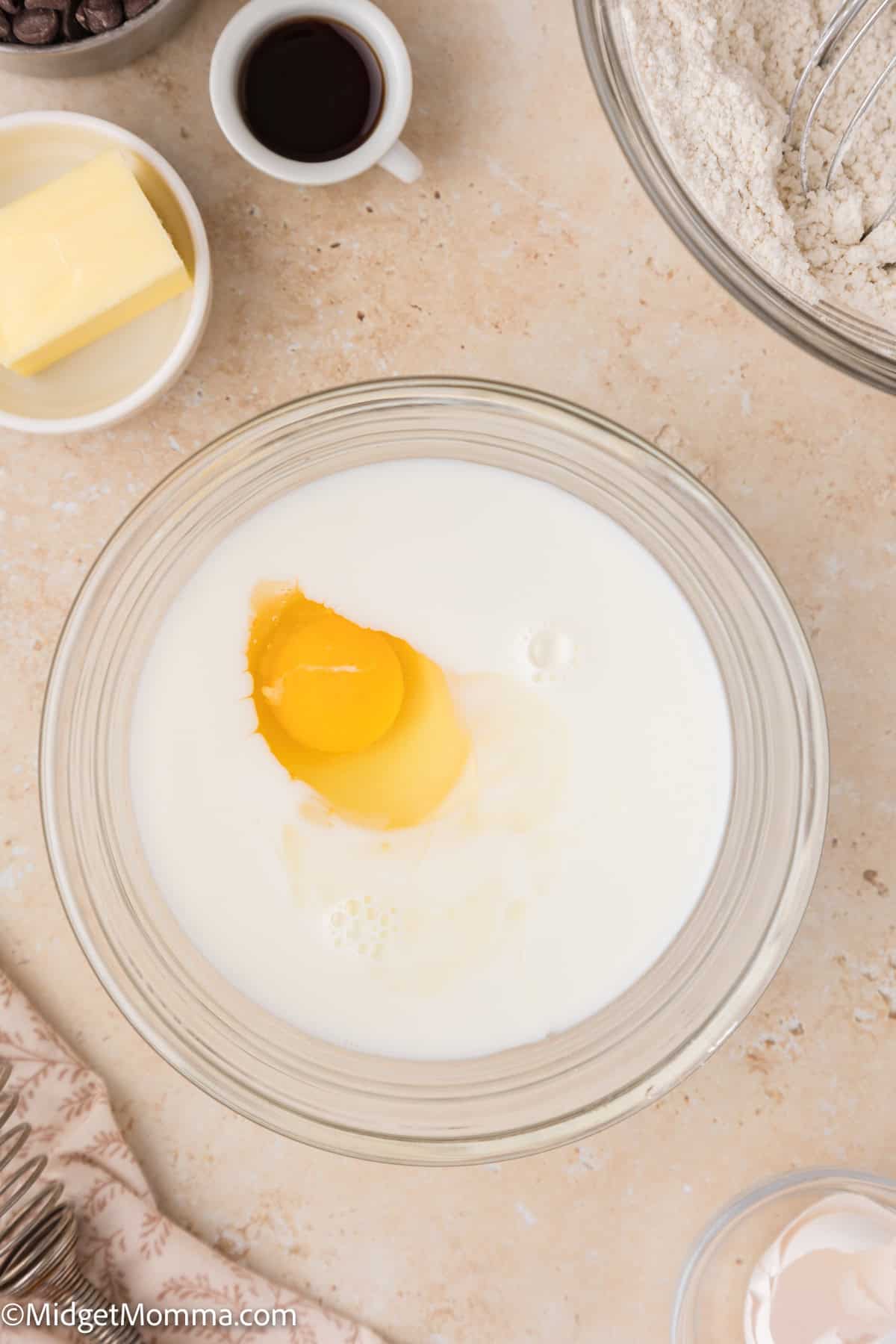 A bowl containing milk and a cracked egg on a counter. Nearby are a cup of vanilla extract, a plate of butter, a bowl of flour, and a small bowl of chocolate chips.