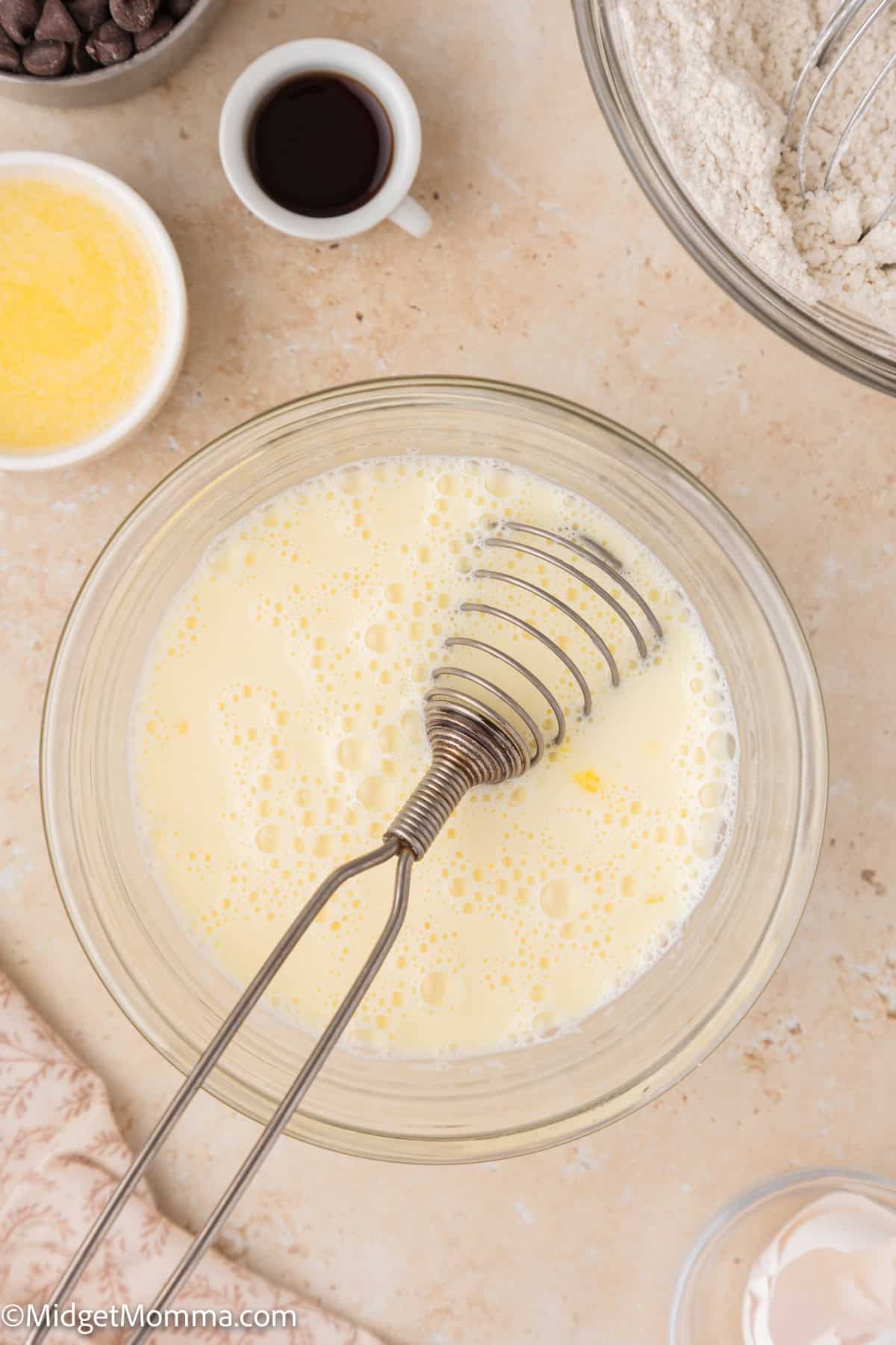 A mixing bowl with whisked eggs and milk, a metal whisk in the bowl. Surrounding the bowl are small containers of chocolate chips, vanilla extract, flour, and melted butter on a countertop.