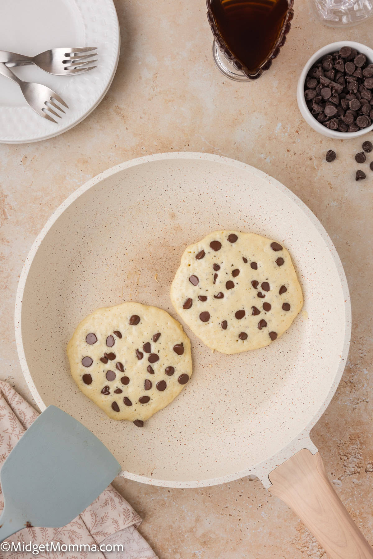 Two chocolate chip pancakes cooking in a skillet. Nearby are a spatula, a plate with forks, a glass of dark liquid, and a bowl of chocolate chips.