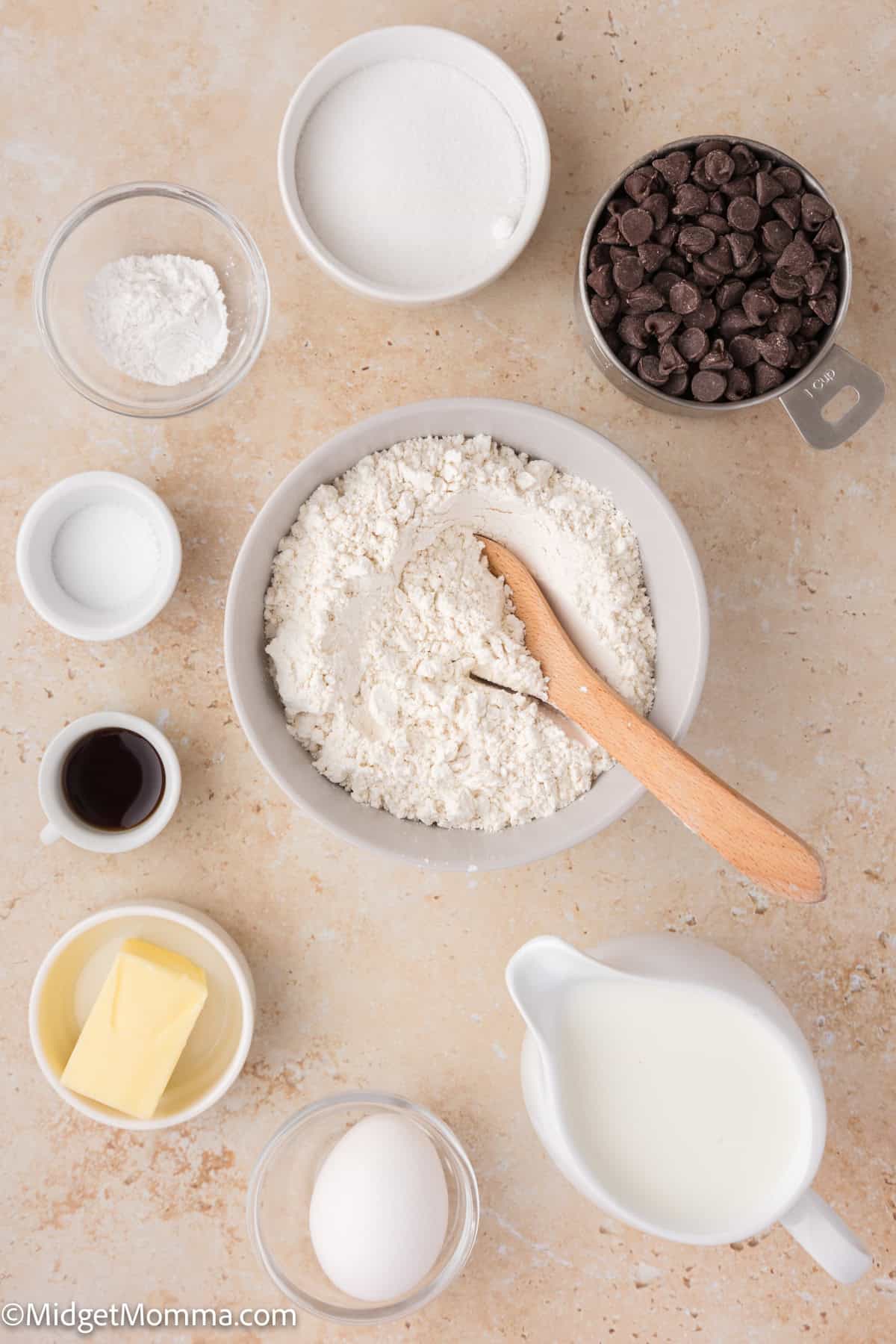 Various baking ingredients including flour, chocolate chips, butter, an egg, sugar, and milk are arranged in bowls on a countertop.