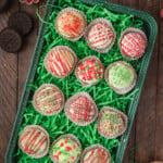 A tray of decorative holiday-themed cake balls with drizzled icing, placed on green shredded paper, surrounded by small bowls of colored sugar and festive sprinkles.