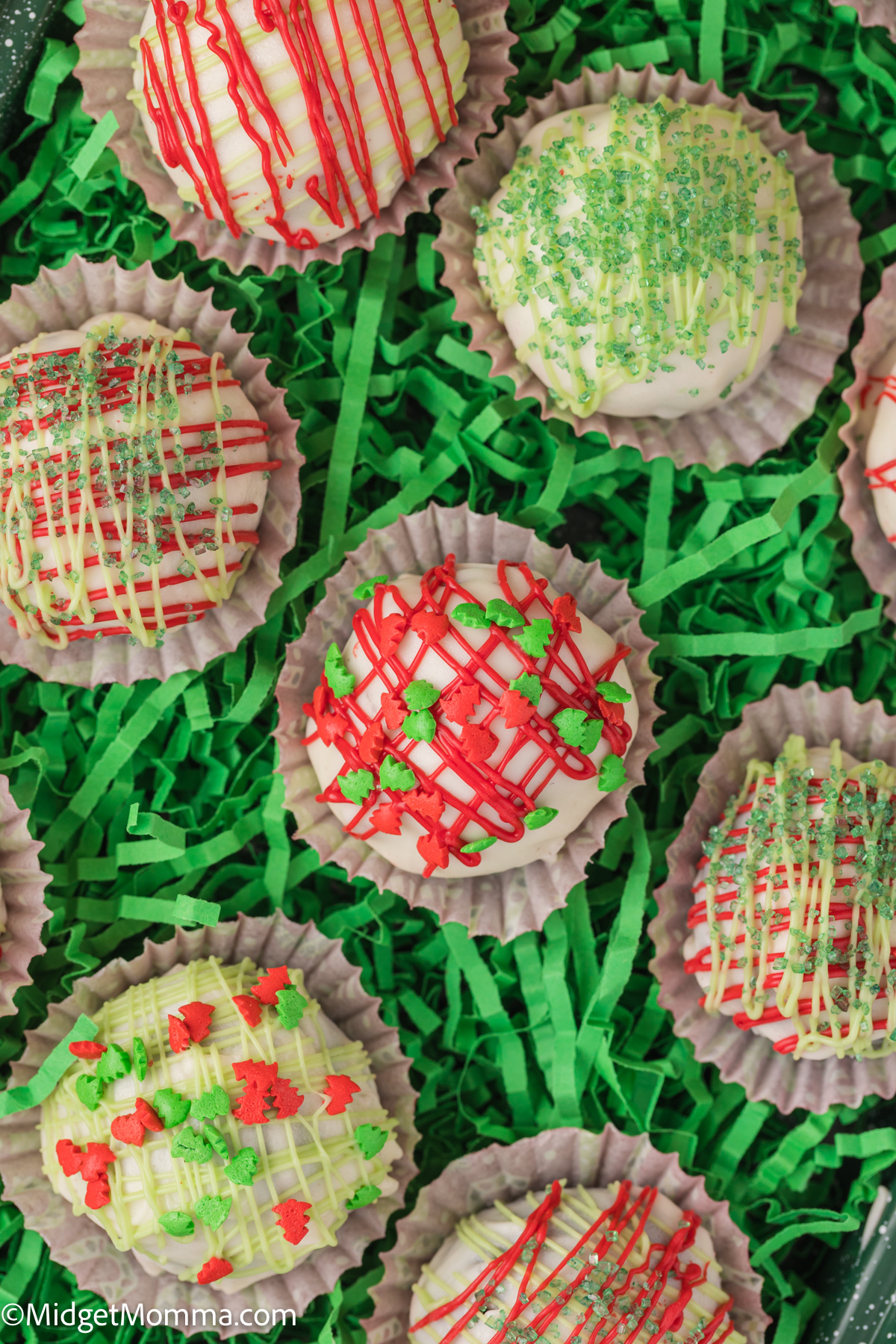 Decorated holiday-themed chocolate-covered Oreo Balls arranged on green shredded paper.