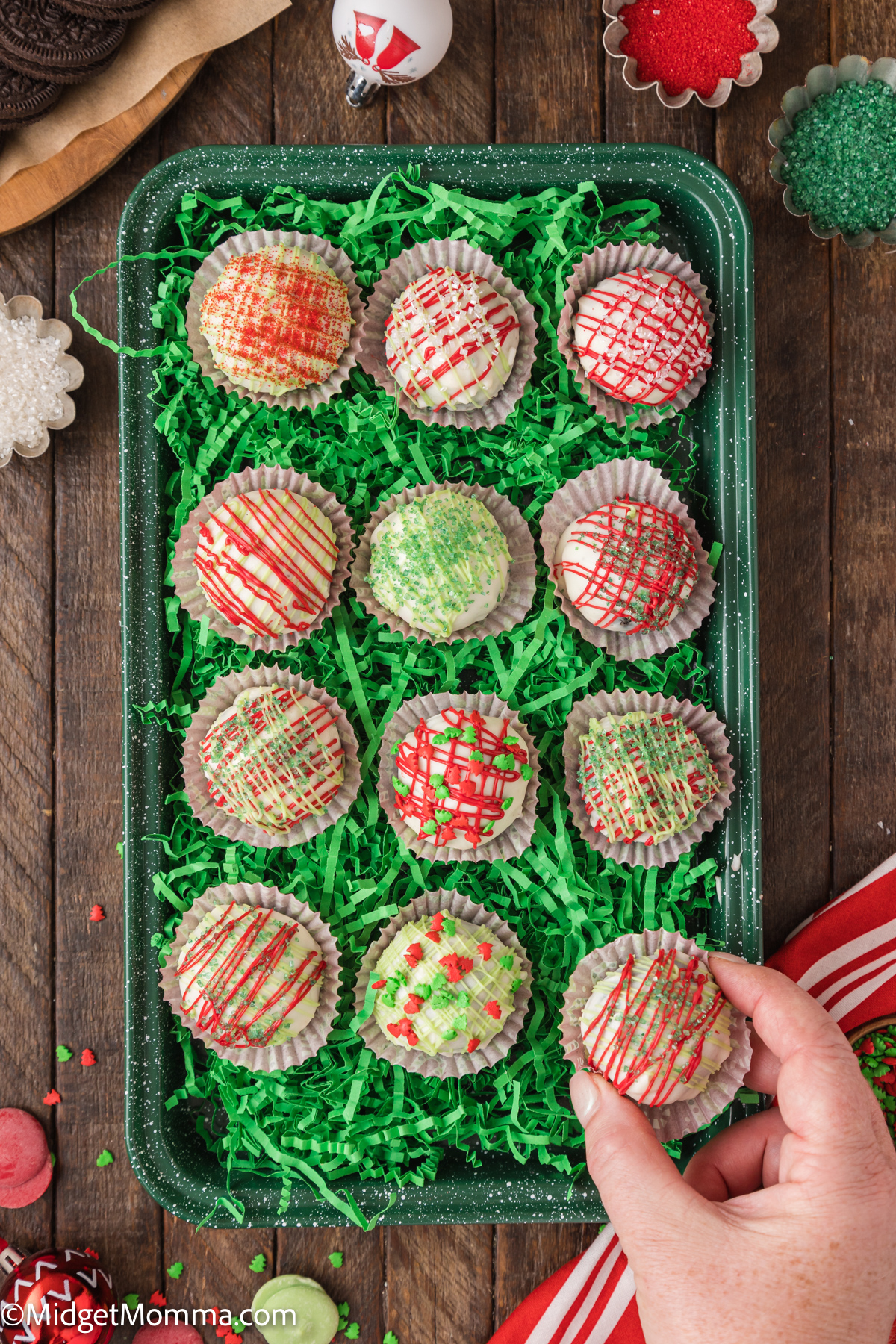 A hand reaching for a decorated chocolate truffle in a tray of colorful, drizzled truffles on a bed of green shredded paper.