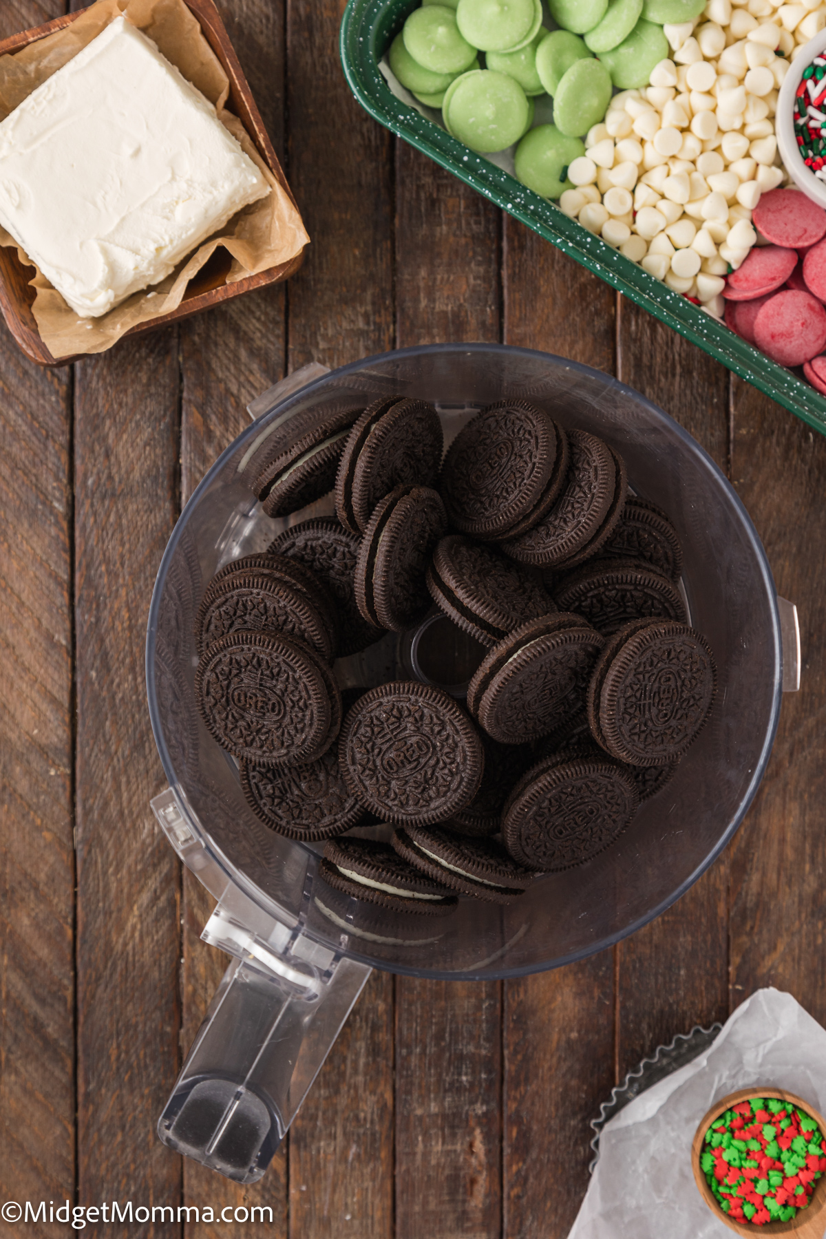 A food processor bowl filled with chocolate sandwich cookies, surrounded by cream cheese, red and green candy melts, white chocolate chips, and sprinkles on a wooden surface.