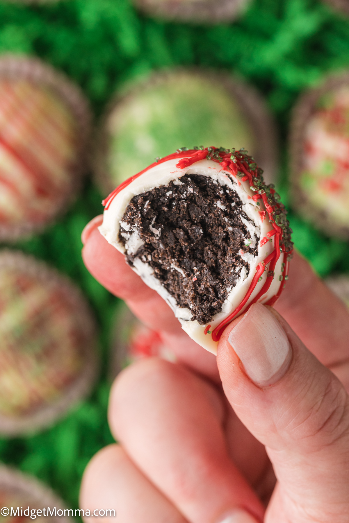 A hand holding a decorative white chocolate-covered oreo balls treat, drizzled with pink icing and sprinkles, with more similar treats on a bed of green grass in the background.