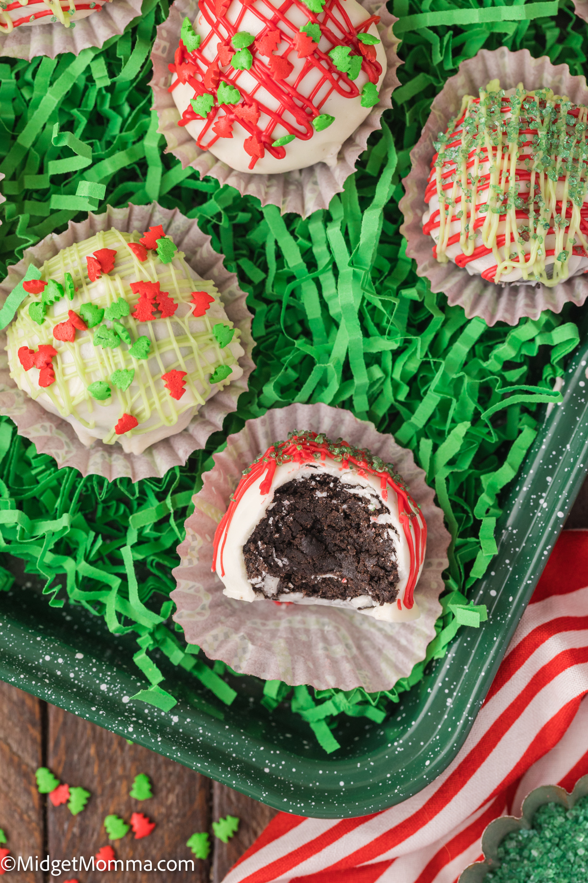 Decorated oreo truffles in paper cups on green shredded paper, featuring colorful icing and sprinkles; one truffle is partially eaten, revealing a dark interior.