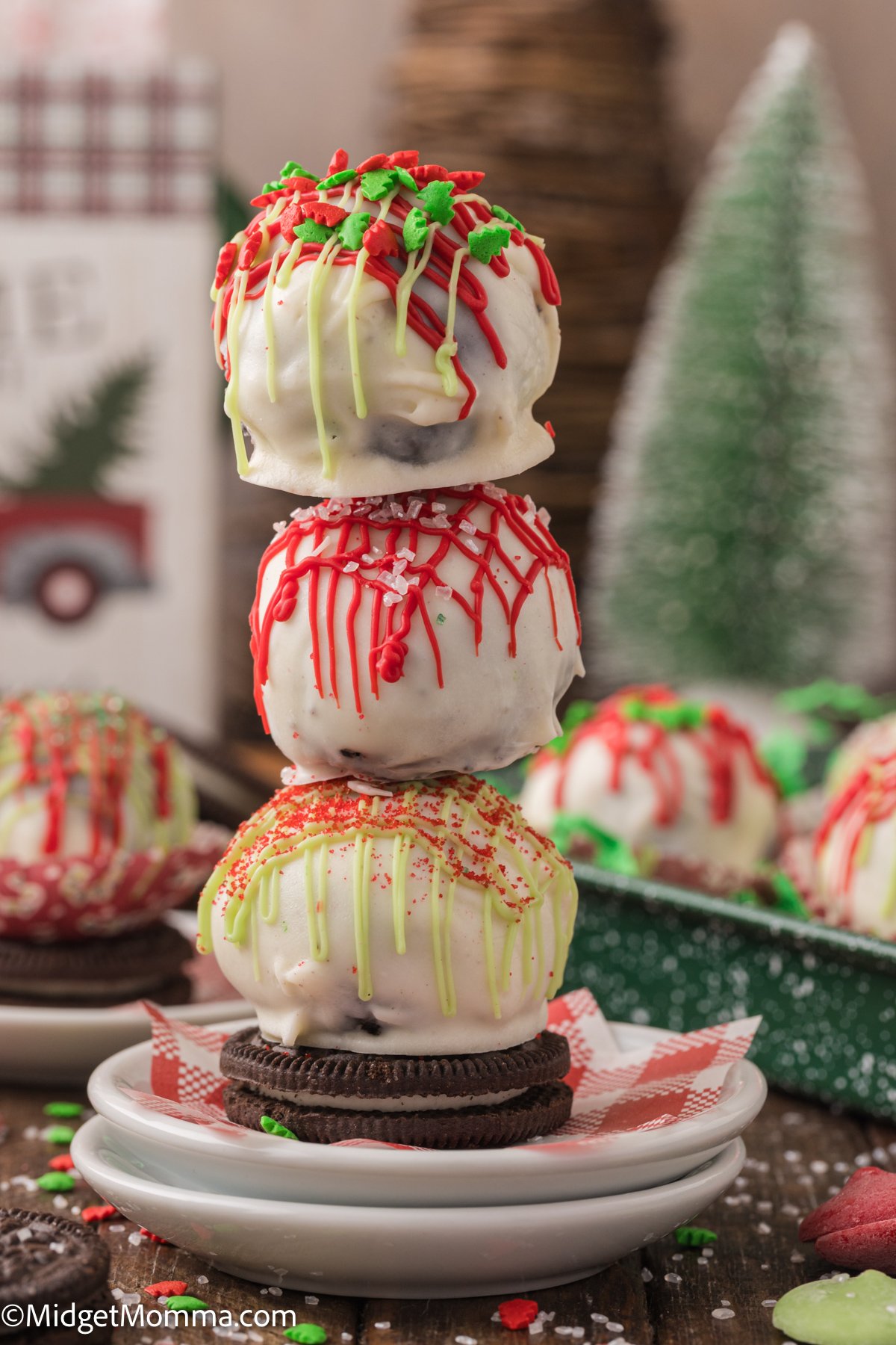 A stack of three festive white chocolate-covered cake balls drizzled with red and yellow icing, topped with sprinkles, placed on a small plate with Oreo cookies. Holiday decorations in the background.