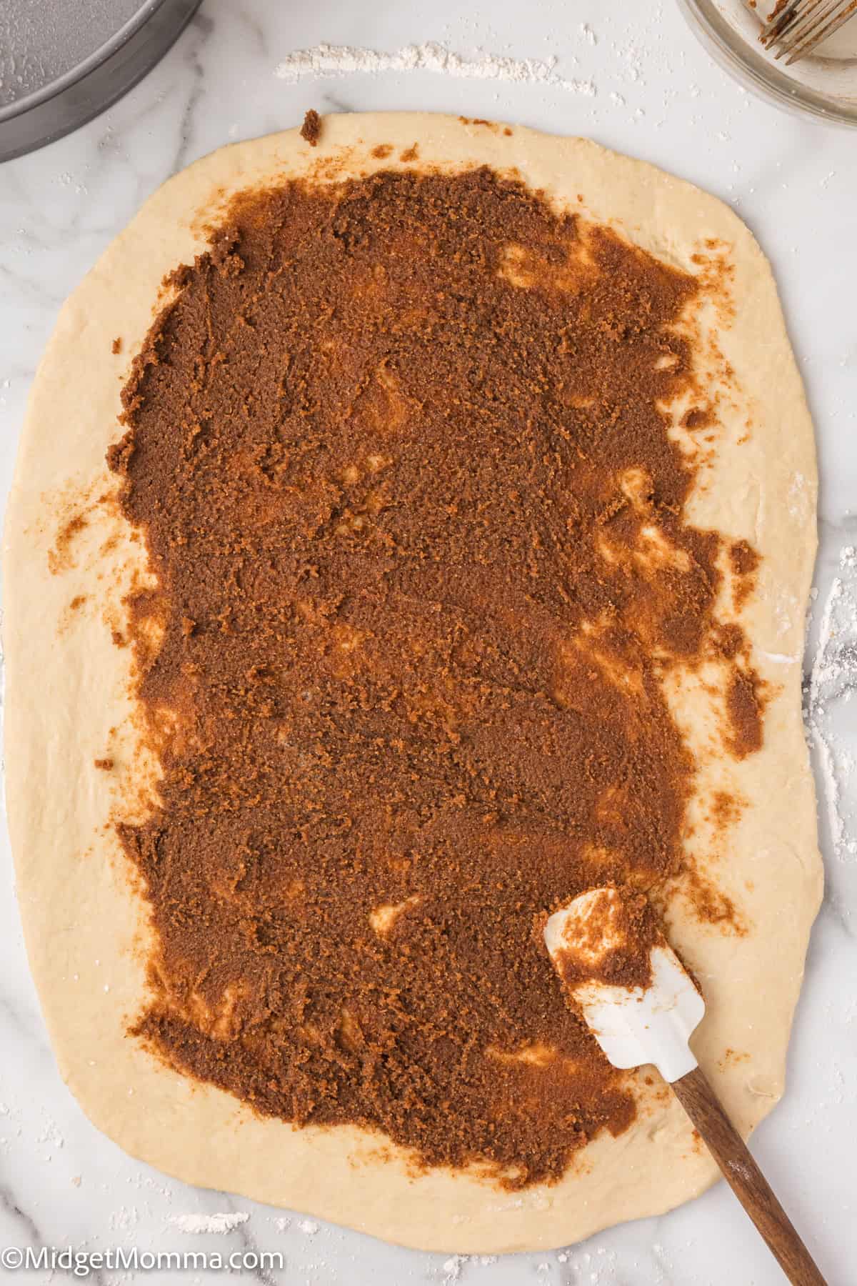 Dough rolled out with brown sugar and cinnamon spread, on a marble surface, next to a spatula.